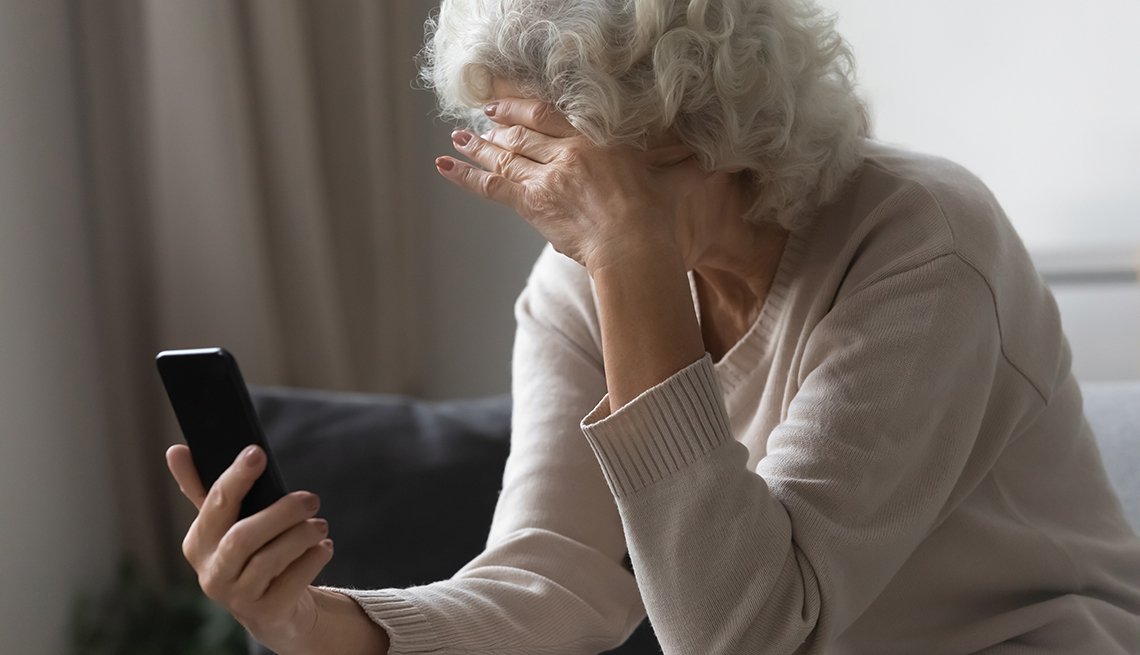 Mujer mayor se cubre su rostro con una mano y con la otra sostiene un teléfono móvil.