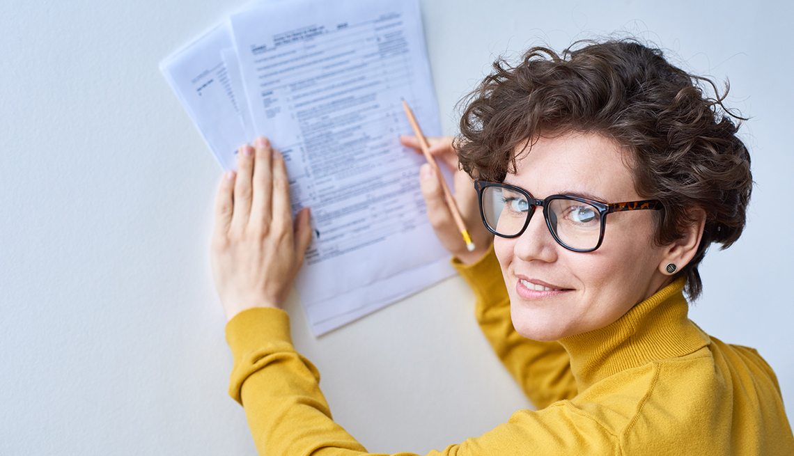 woman wearing yellow turtleneck sweater and glasses looks confident while using a wall to support tax forms she is holding and filling out