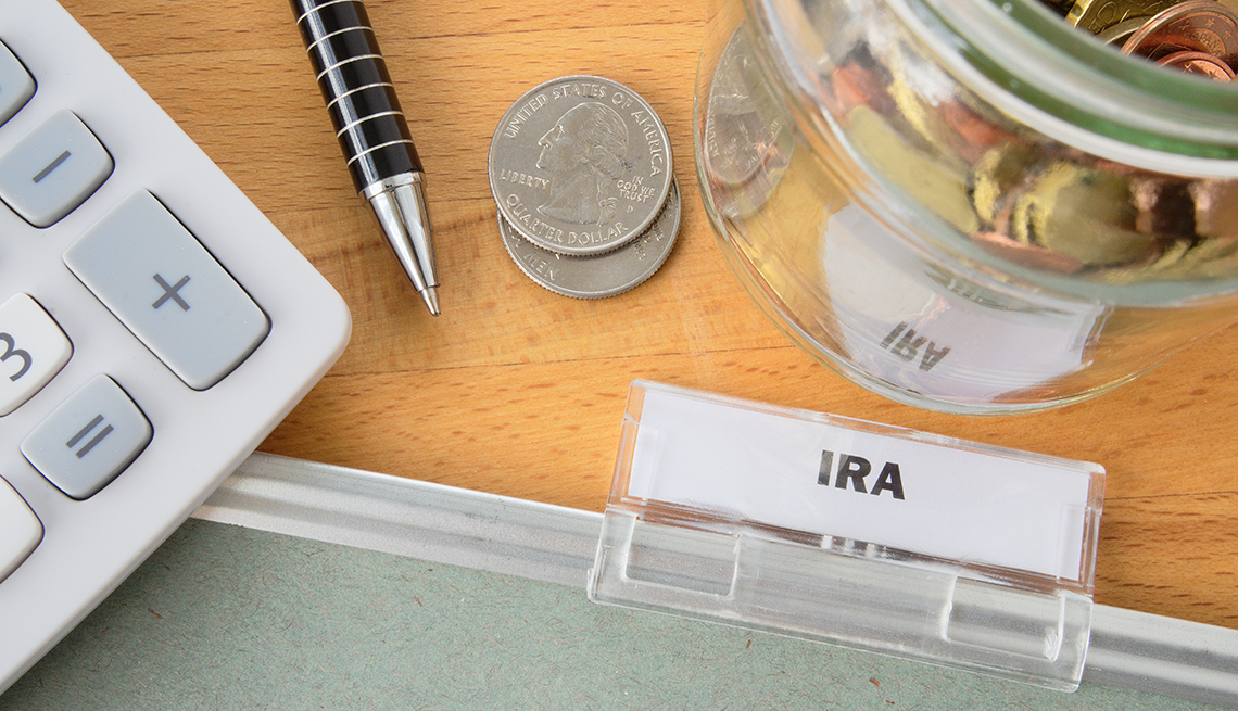 Visualización en el escritorio de una pestaña de carpeta de archivos en papel con la etiqueta "IRA" junto con un frasco de dinero, veinticinco centavos, un bolígrafo y una calculadora.