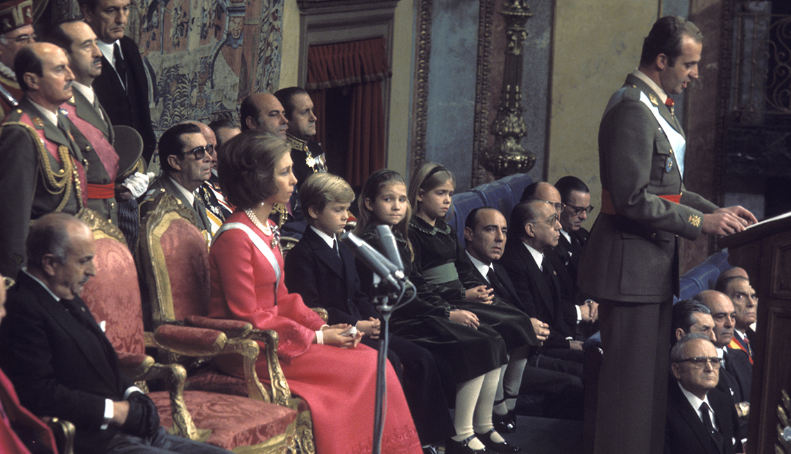 Foto de la coronación del rey Juan Carlos, mientras el da un discurso y la reina Sofia y sus hijos sentados detrás de él, al lado de altos oficiales del gobierno.