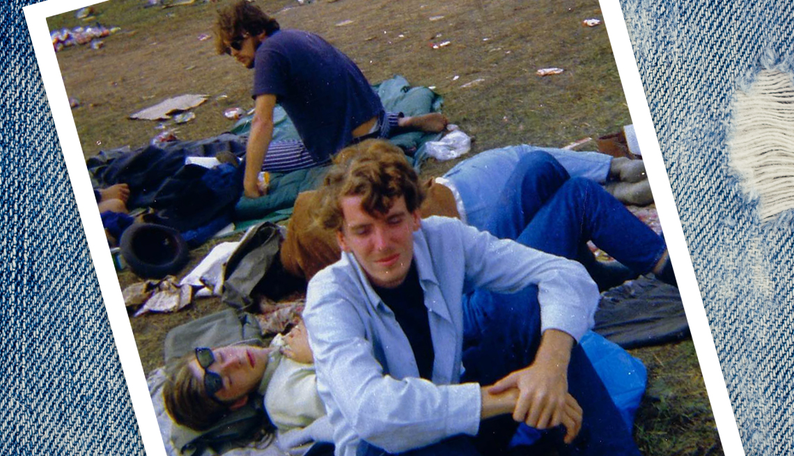 item 3 of Gallery image - young man wearing bell bottom jeans sitting on a sleeping bag on the field at Woodstock, next to a group of people sleeping and lying on the ground.