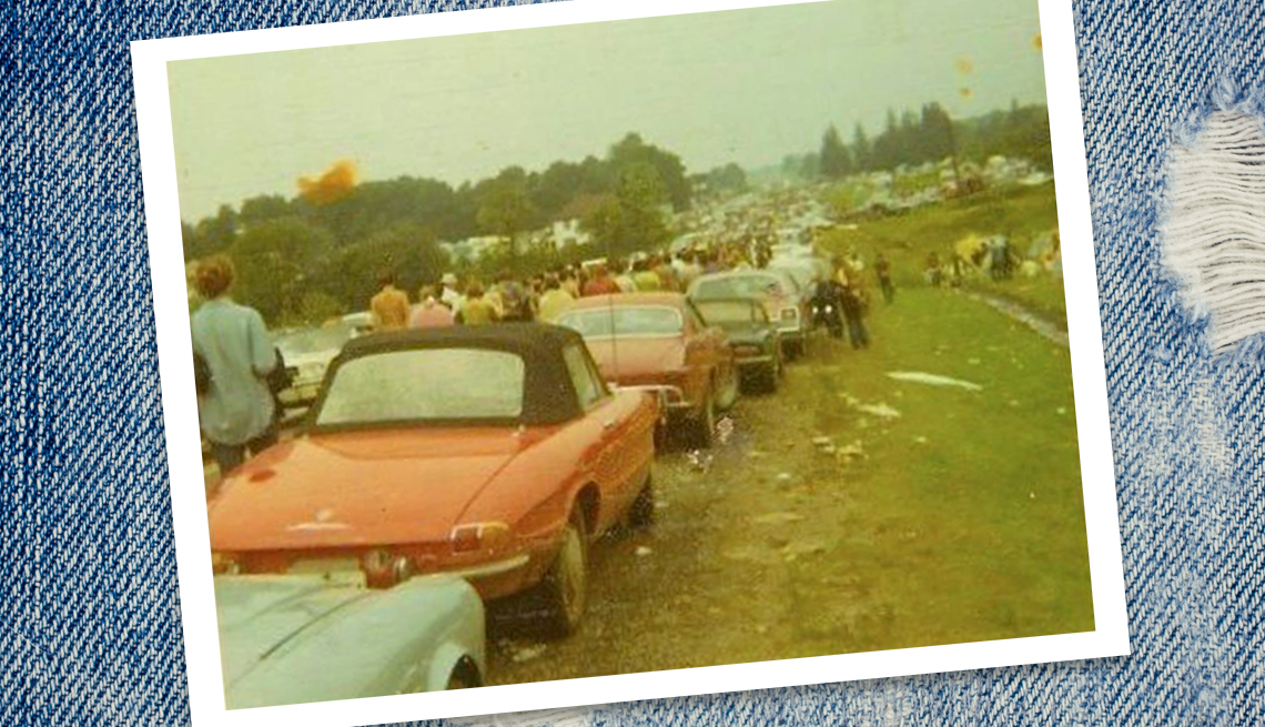 a long line of abandoned cars stopped in the field leading up to Woodstock, with people walking past them towards the festival barely visible in the distance