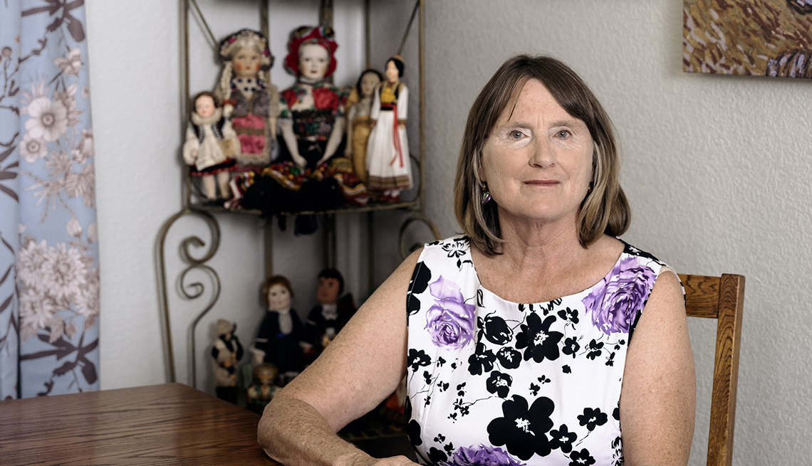 claudia oreilly sits at the table with a collection of dolls behind her on a shelf