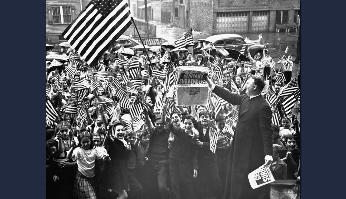 item 5 of Gallery image a priest holds up a newspaper announcing the end of the war in front of a crowd of schoolboys waving american flags
