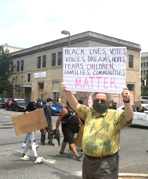 photo of man at protest wearing covid face mask and holding up a sign that reads black lives votes voices dreams hopes fears children families communities matter