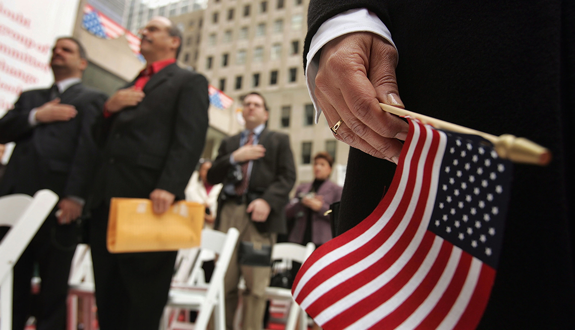 Mano sosteniendo una bandera americana con personas detrás jurando como ciudadanos