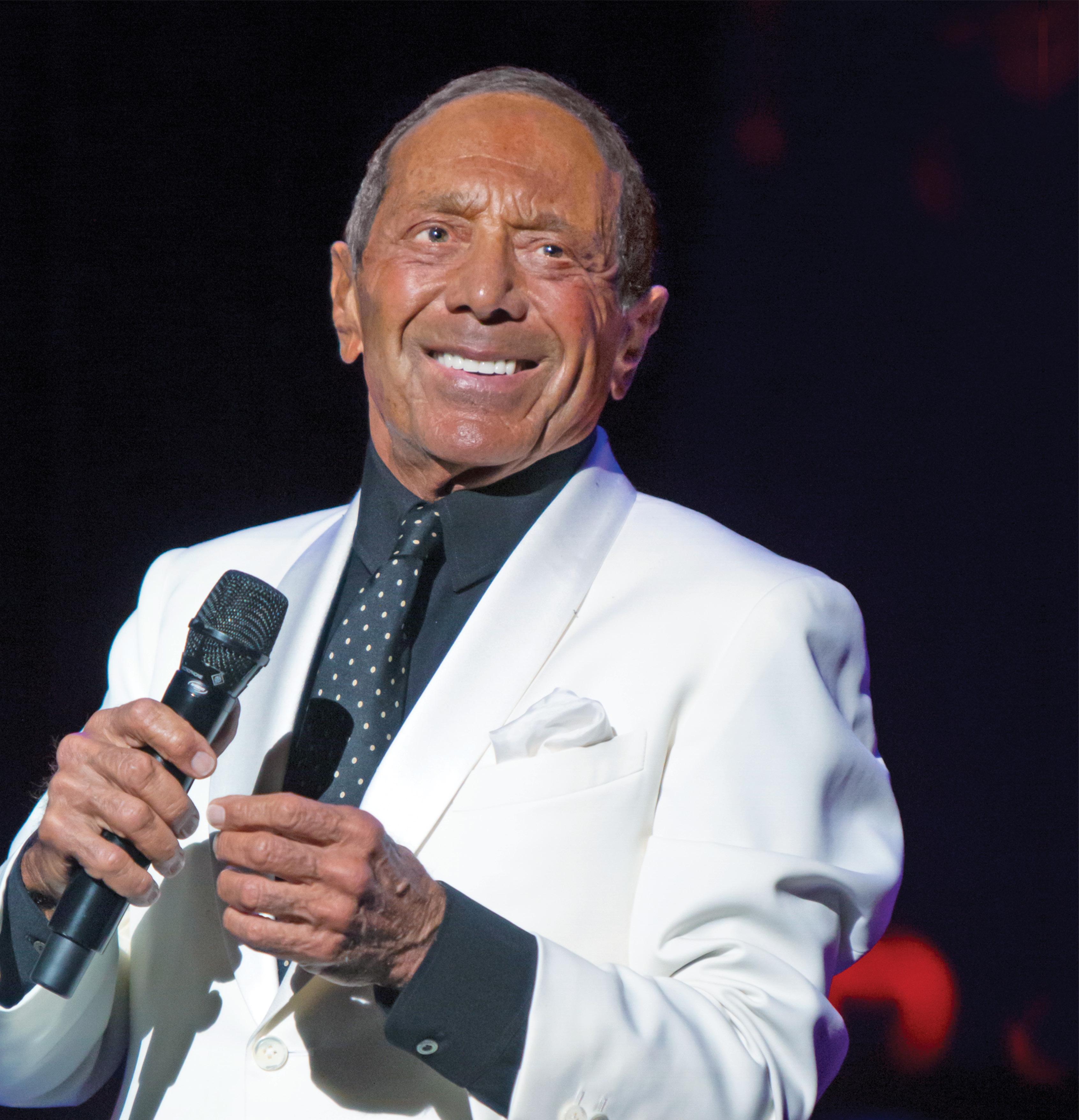 Photograph of Paul Anna in a white suit holding a mic and smiling