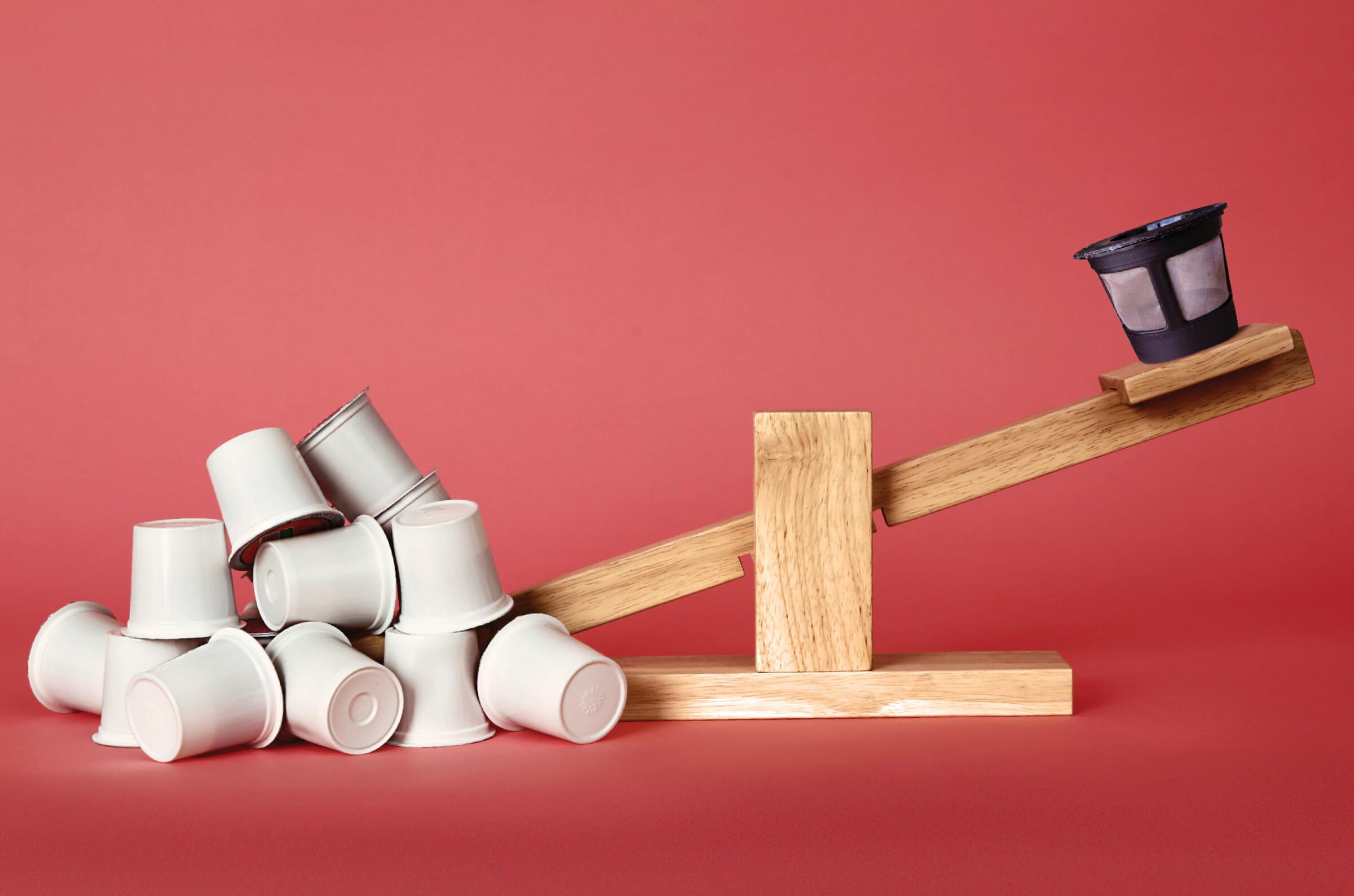 Simple wooden scale with one end piled with disposable K-cups weighing down the other end holding one reusable K-cup.
