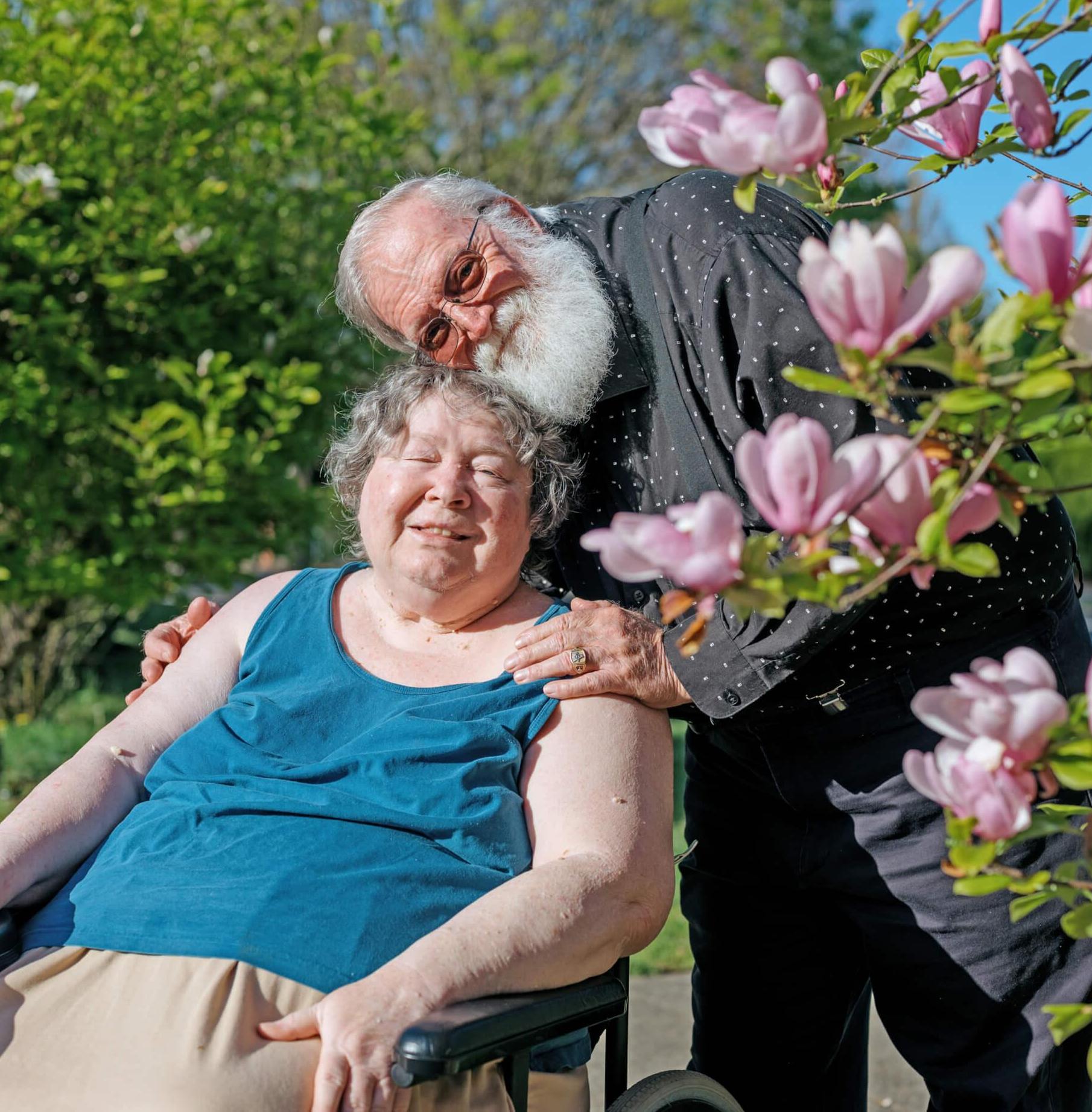 An white haired caucasian woman, Susan Bliven, in a wheelchair outside with her husband Lee standing behind her with his hands on her shoulders.