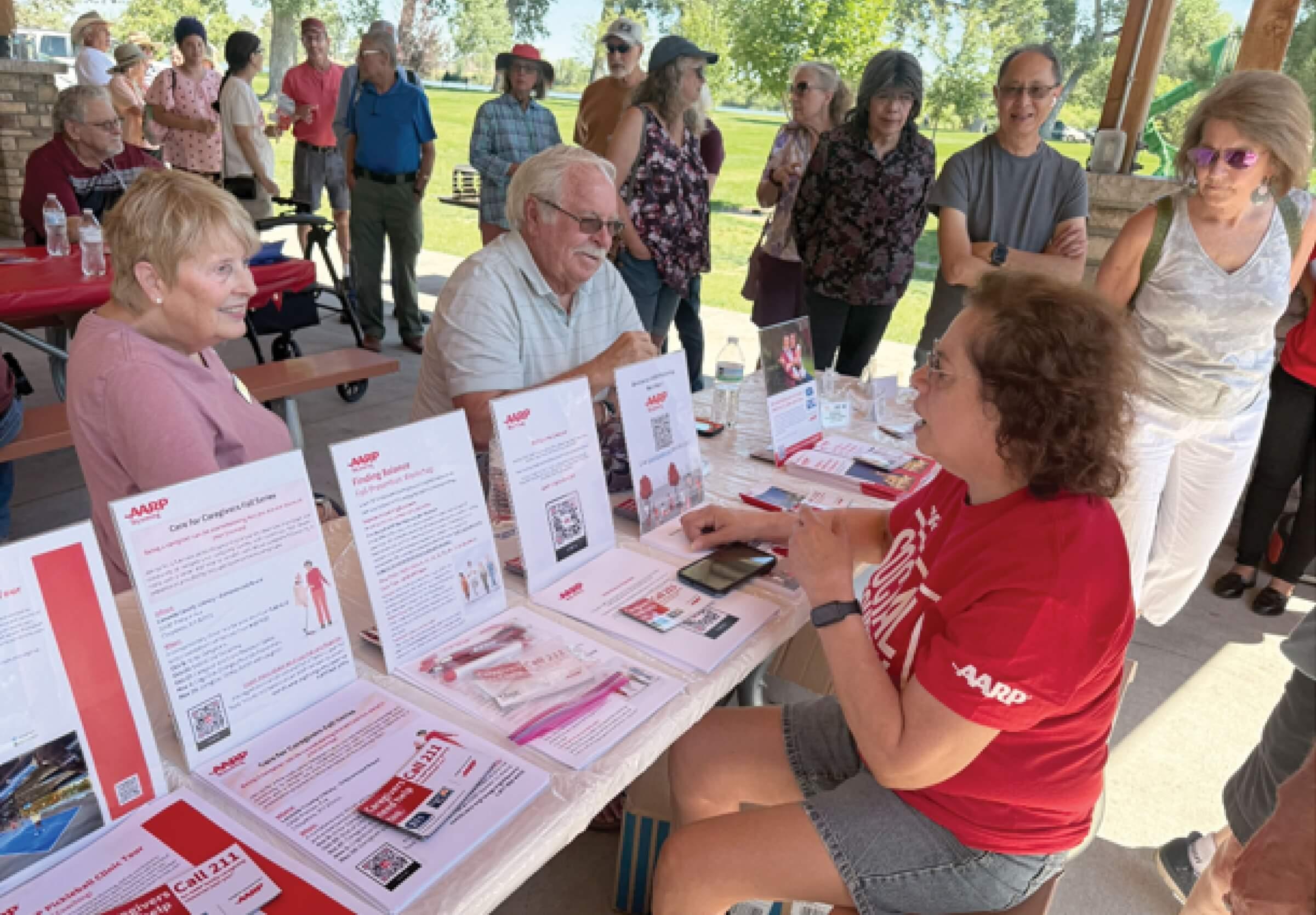 Photograph of Alice Swank sitting at an AARP Social Security picnic. Two people sit in front of her while a line of people is standing to the side waiting to speak to her.