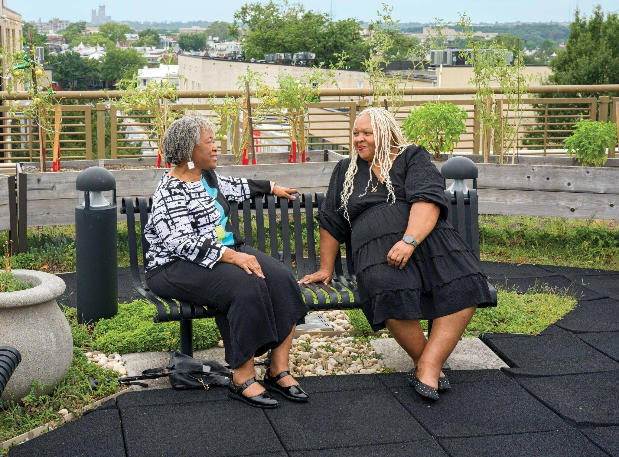 Photograph of Angela Jasper in a skirt and black and white top sitting on a bench and talking to visitor Robin Baxter, who is wearing a long black dress