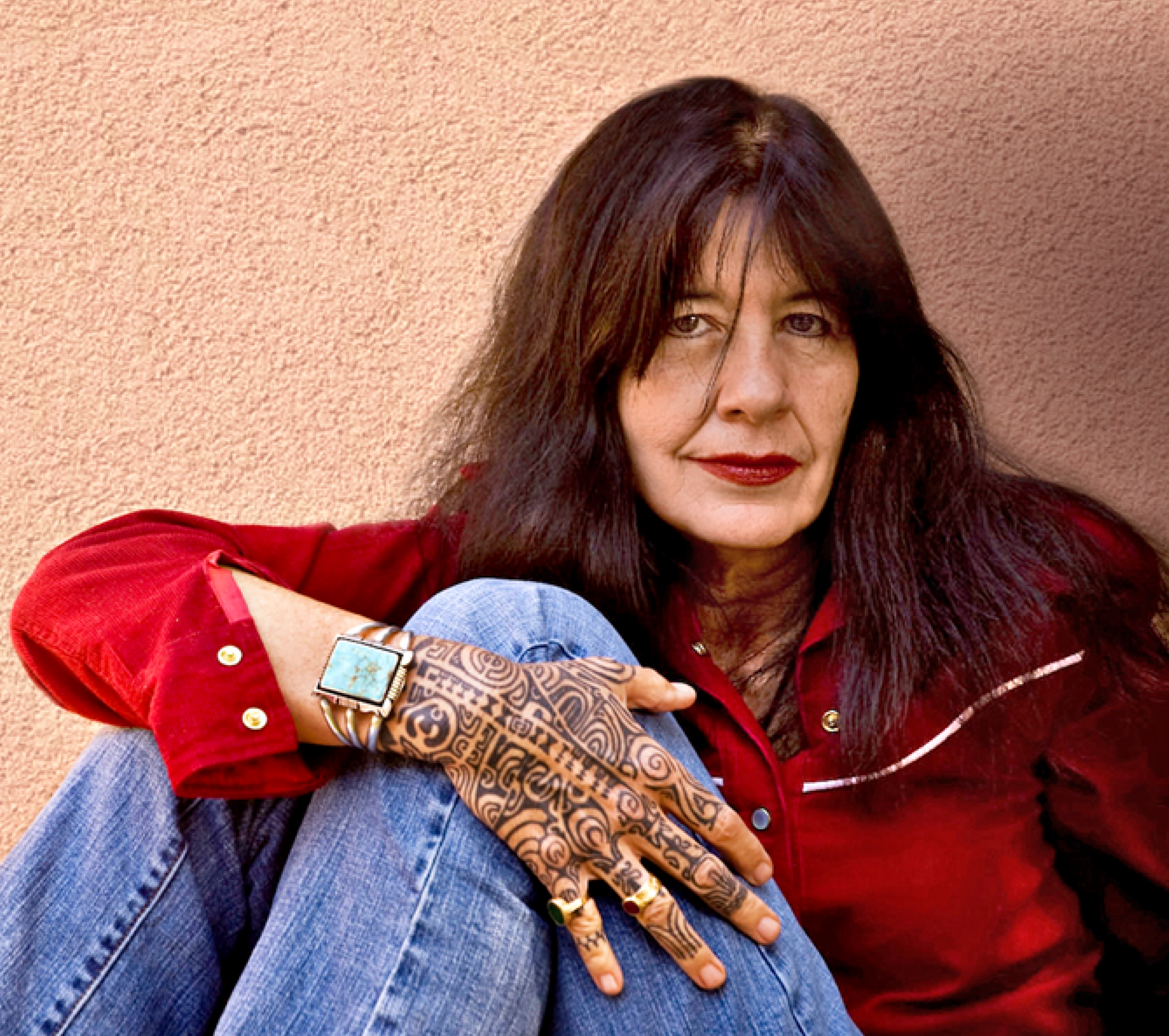 Portrait of Joy Harjo. She is wearing her hair down, a red blouse and jeans.