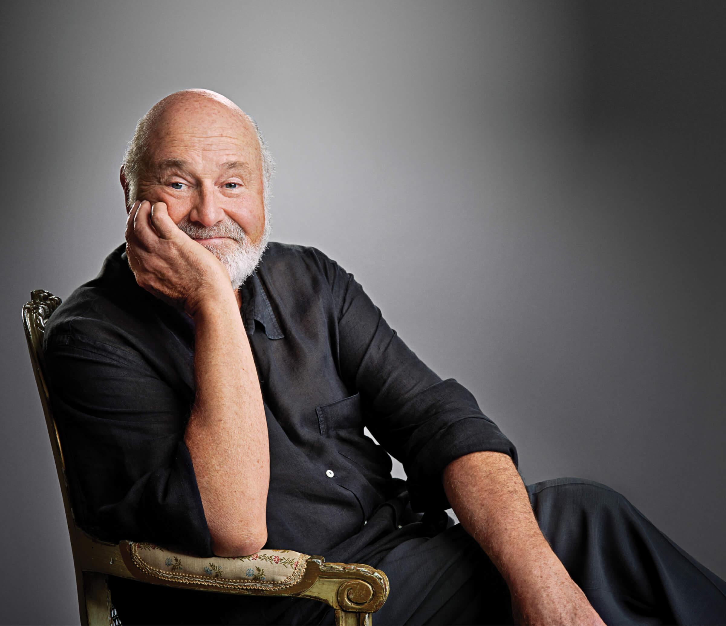 Rob Reiner sitting on a chair and simling at the camera.