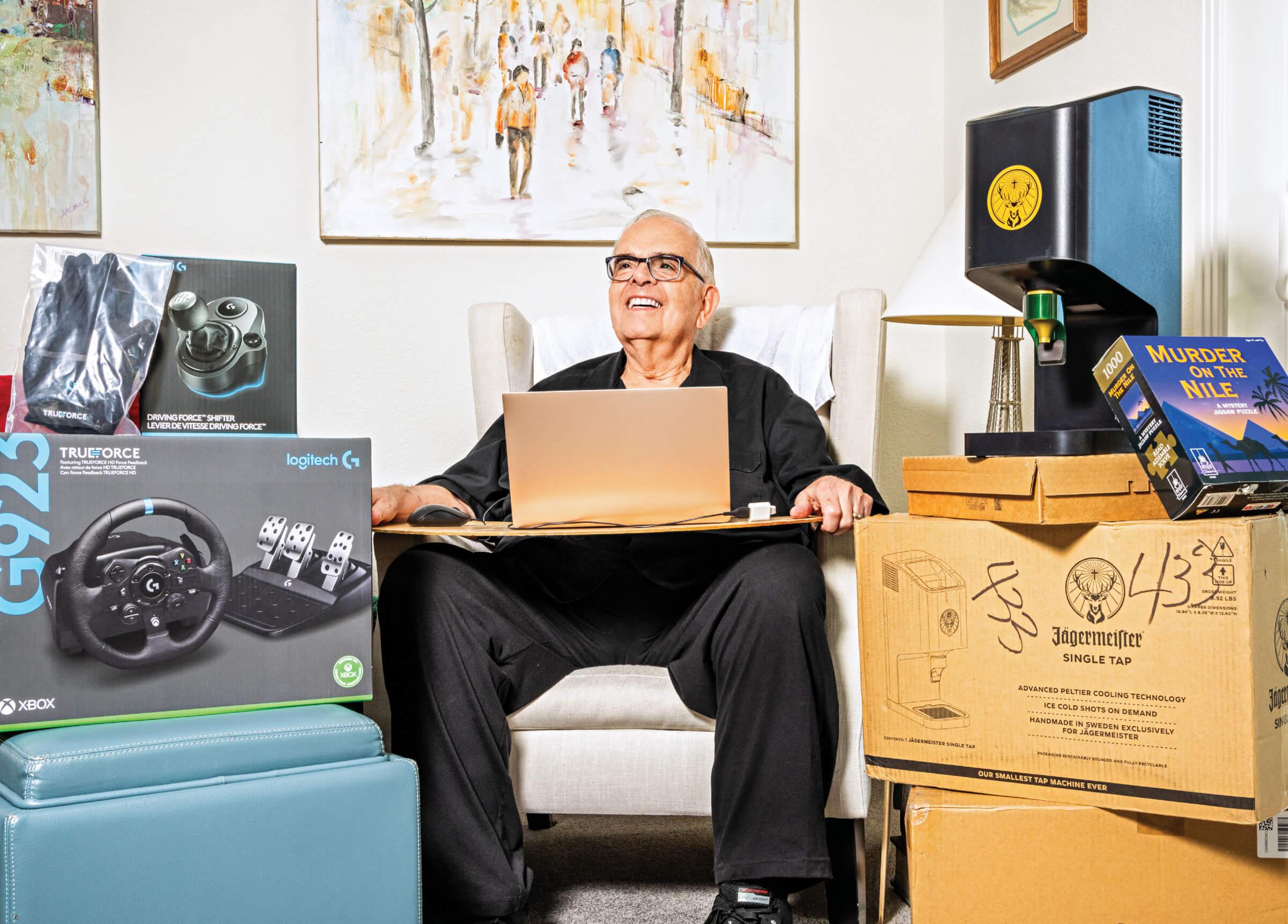 Image of Steve d'Adolf sitting on a couch with a laptop on this lap. He is surrounded by several boxes of different types of electronics.