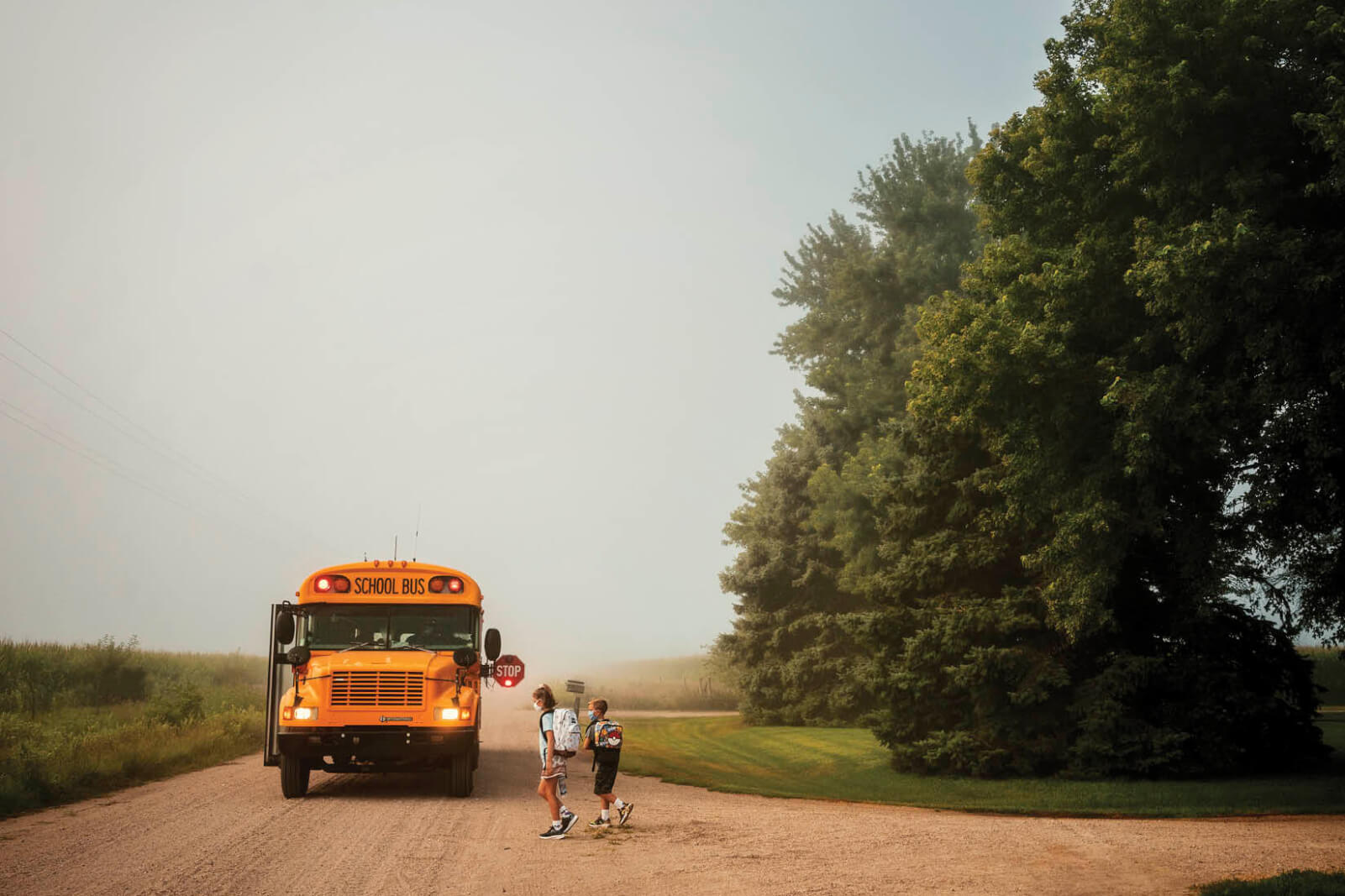 Photo of 2 children on a country road waiting for an approaching yellow school bus