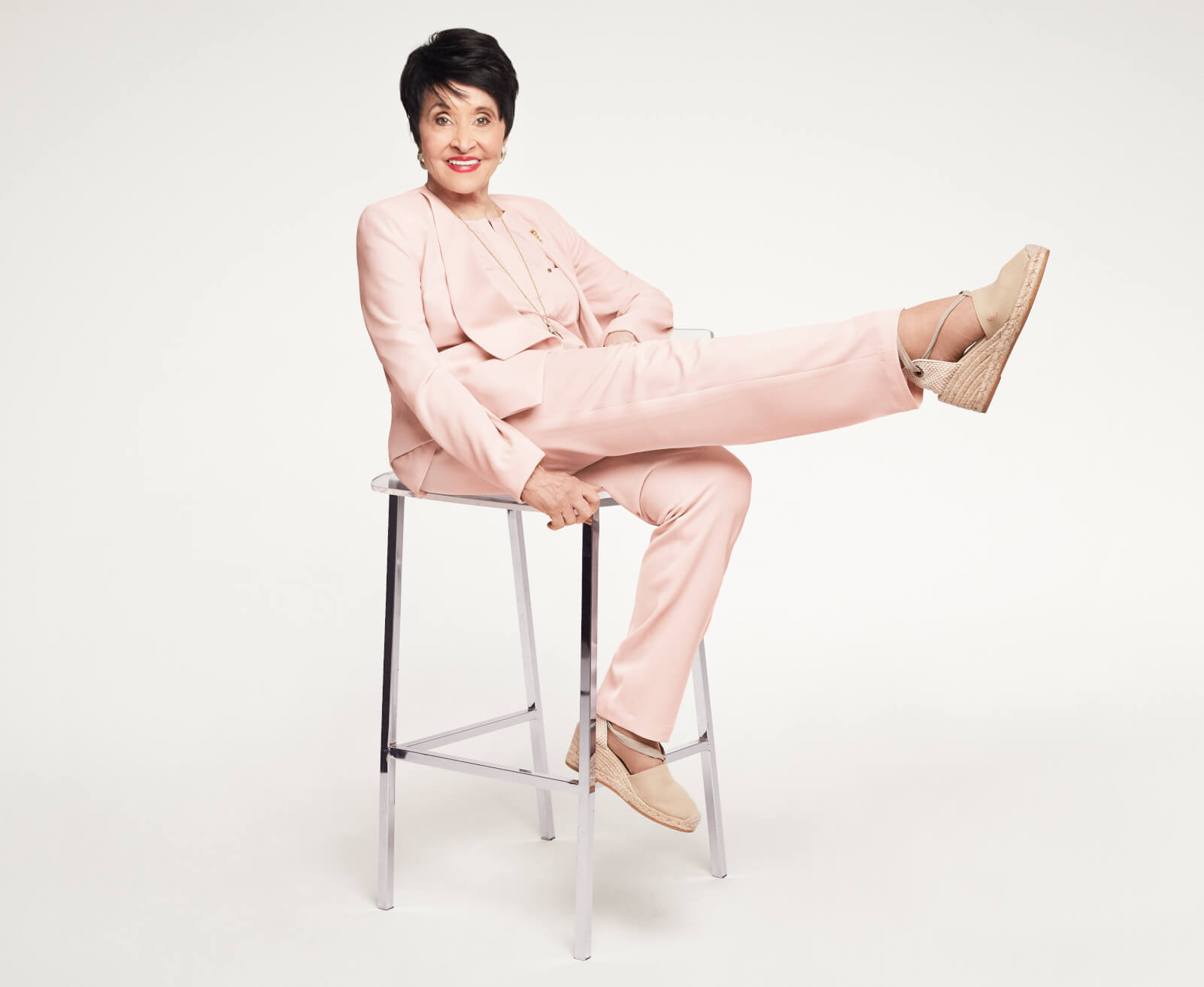 Photo of Chita Rivera wearing a pink suit sitting on a stool with her right leg stretched out horizontally