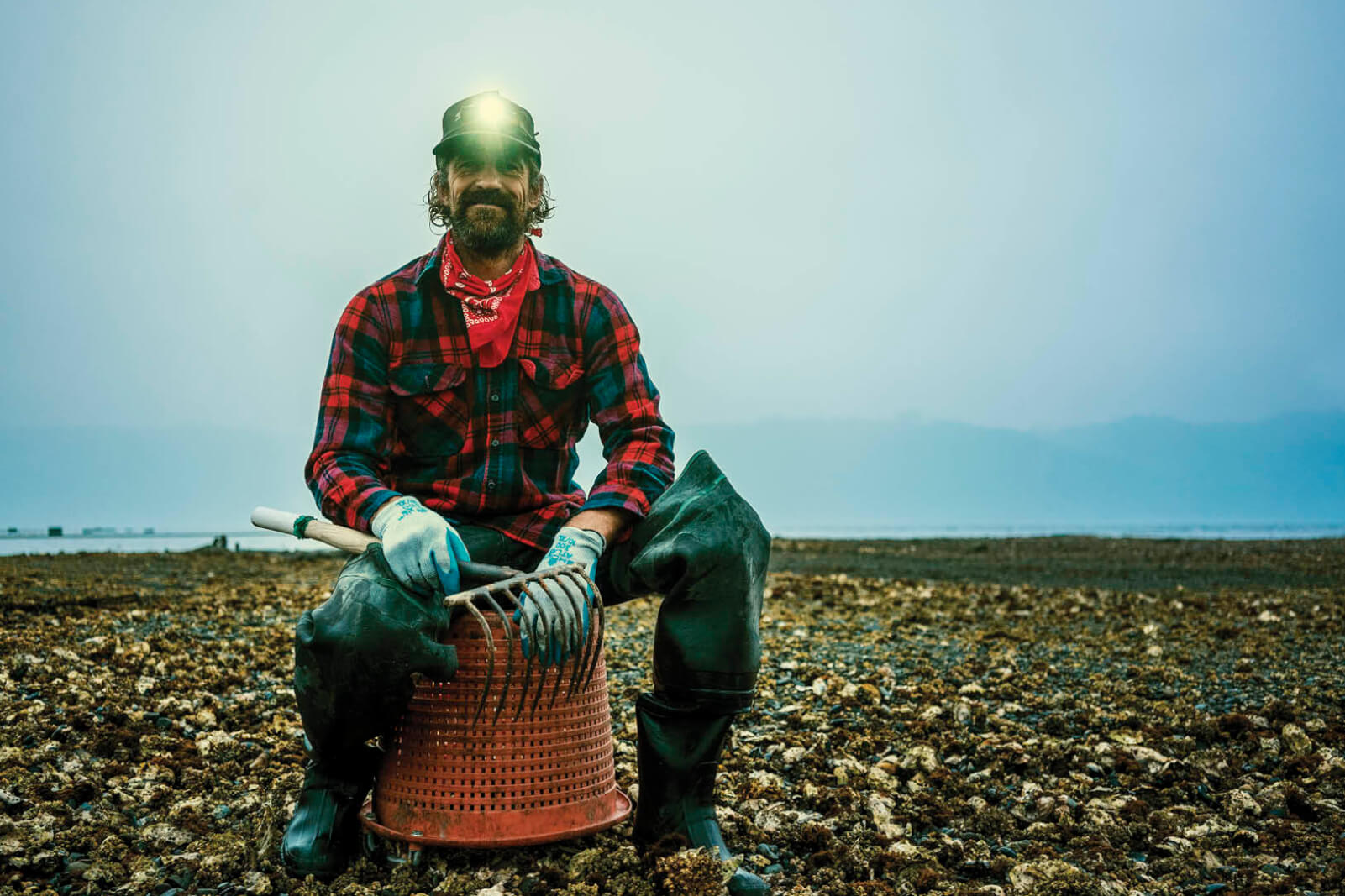 Photo of a man sitting on a large bucket in the middle of a large oyster bed