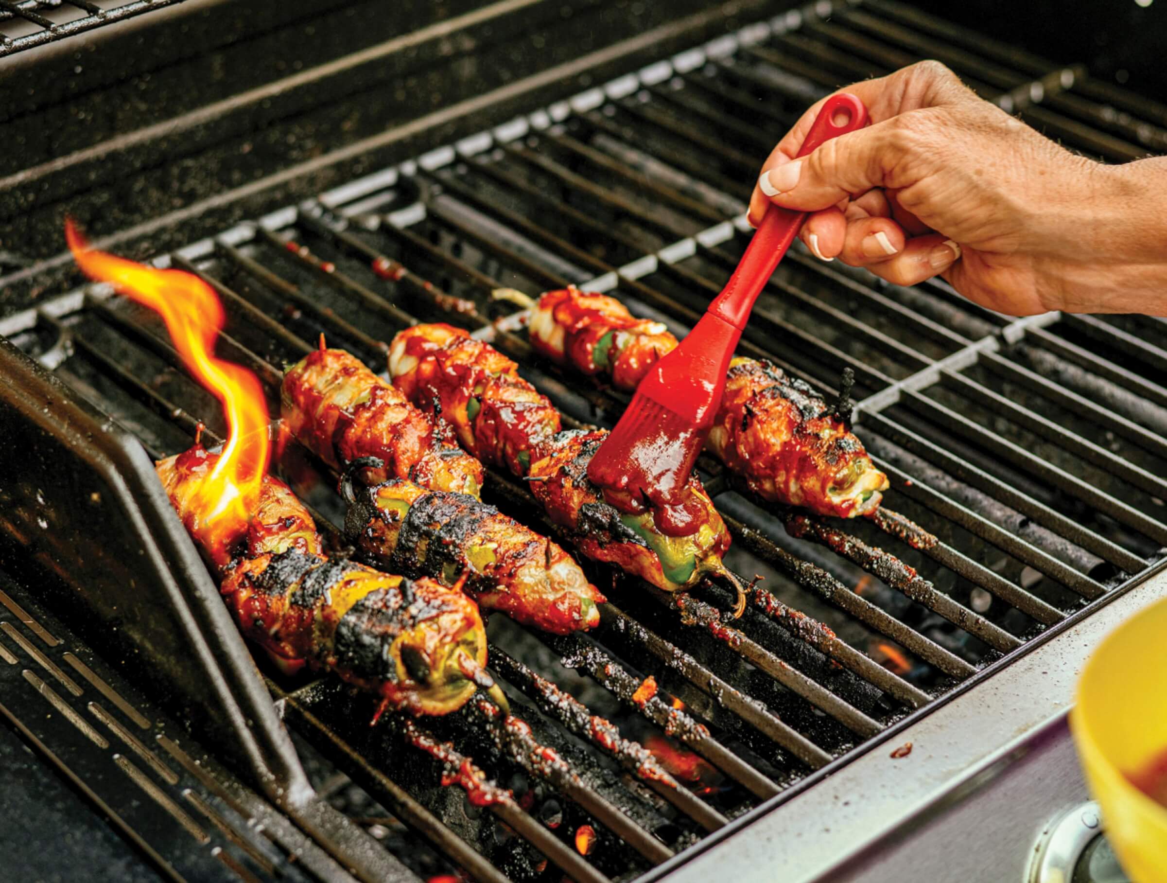 Photo of someone brushing barbecue sauce on skewered peppers cooking on an outdoor grill