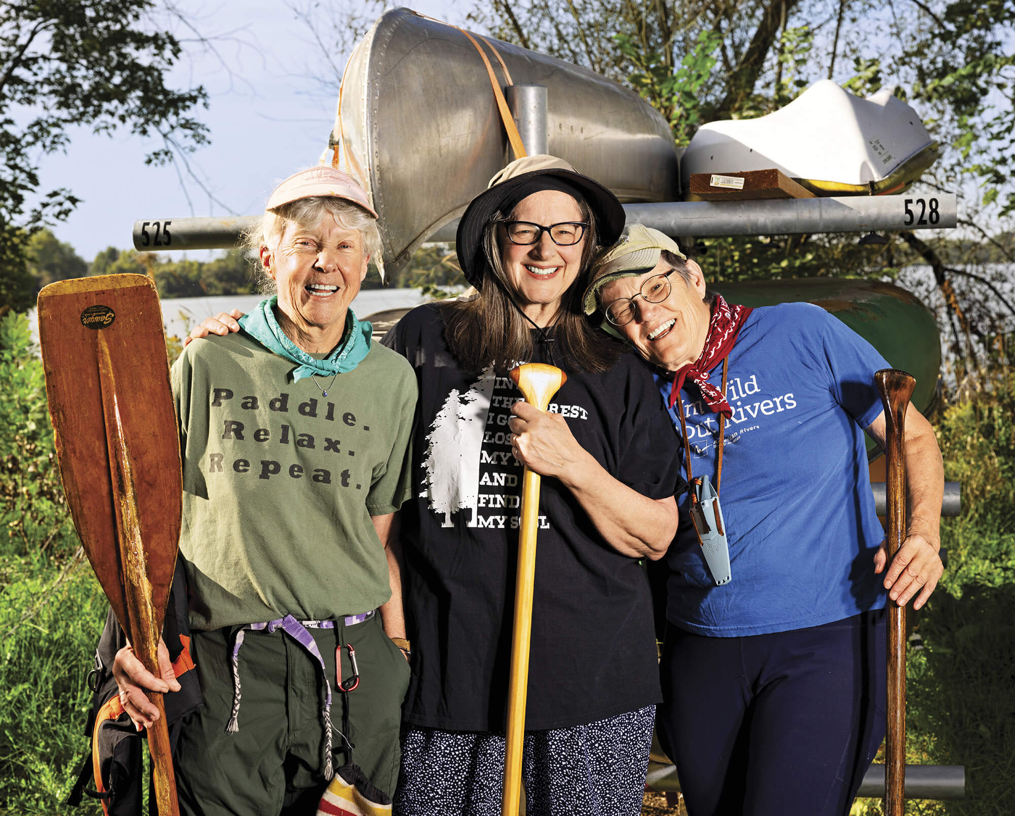 Photo portrait of three women smiling and holding paddles in front of canoes on a rack