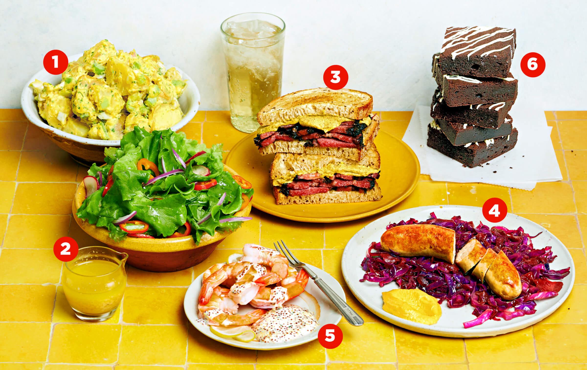 Photo of spread of food on counter: Potato salad, salad, pastrami sandwich, brats and cabbage, shrimp, and brownies