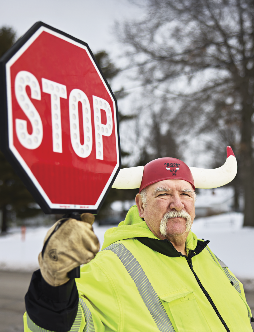 Image of Al Novreske standing on the road holding a stop sign in the cold. He is wearing a hat with horns that says Chicago Bulls on it