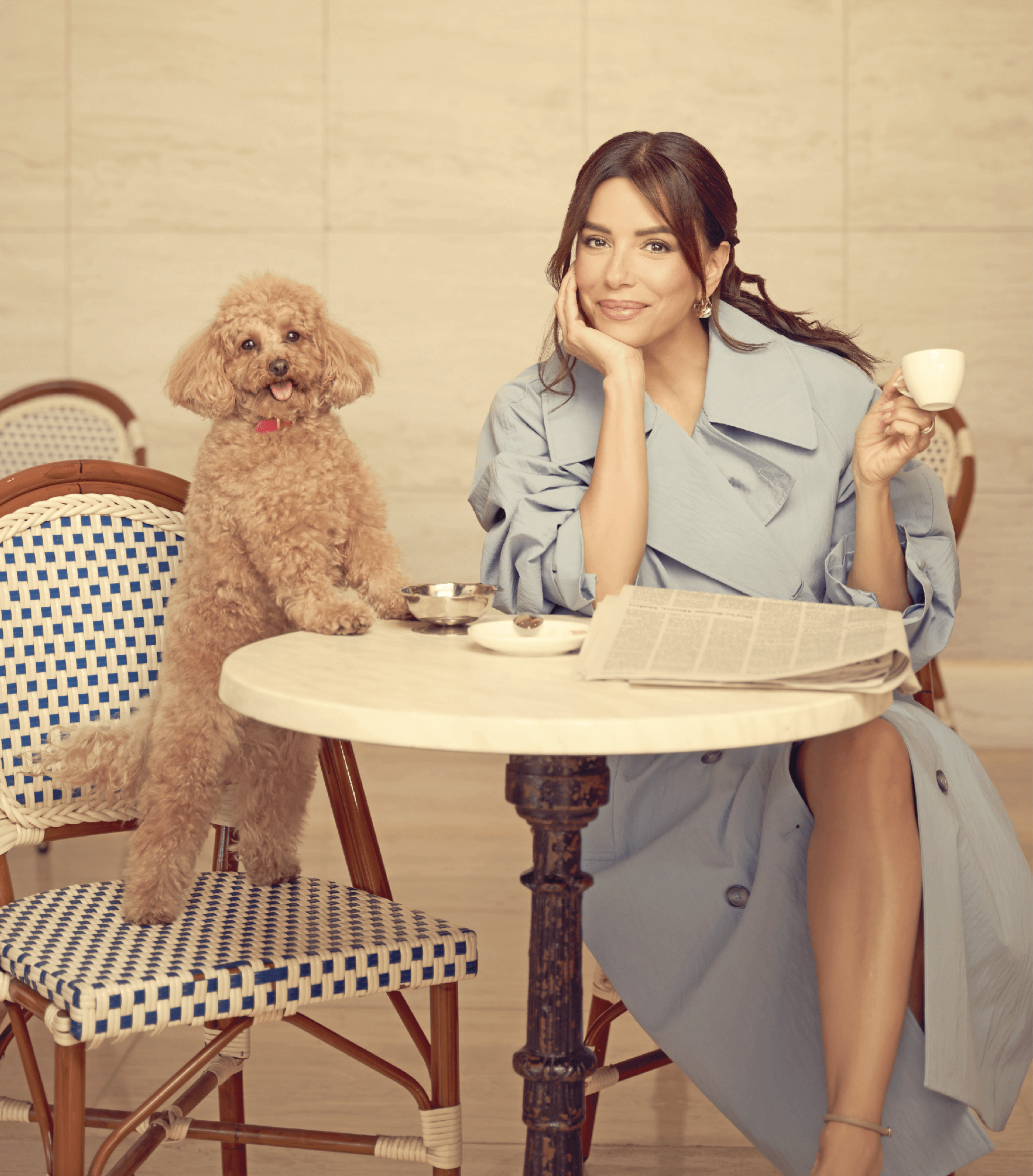 Image of Eva Longoria sitting at a table and holding a small coffee cup in one hand. Her dog is sitting on the other chair smiling into the camera. Eva is wearing a light blue trench coat dress and has her hair open. A newspaper lies open on the table.