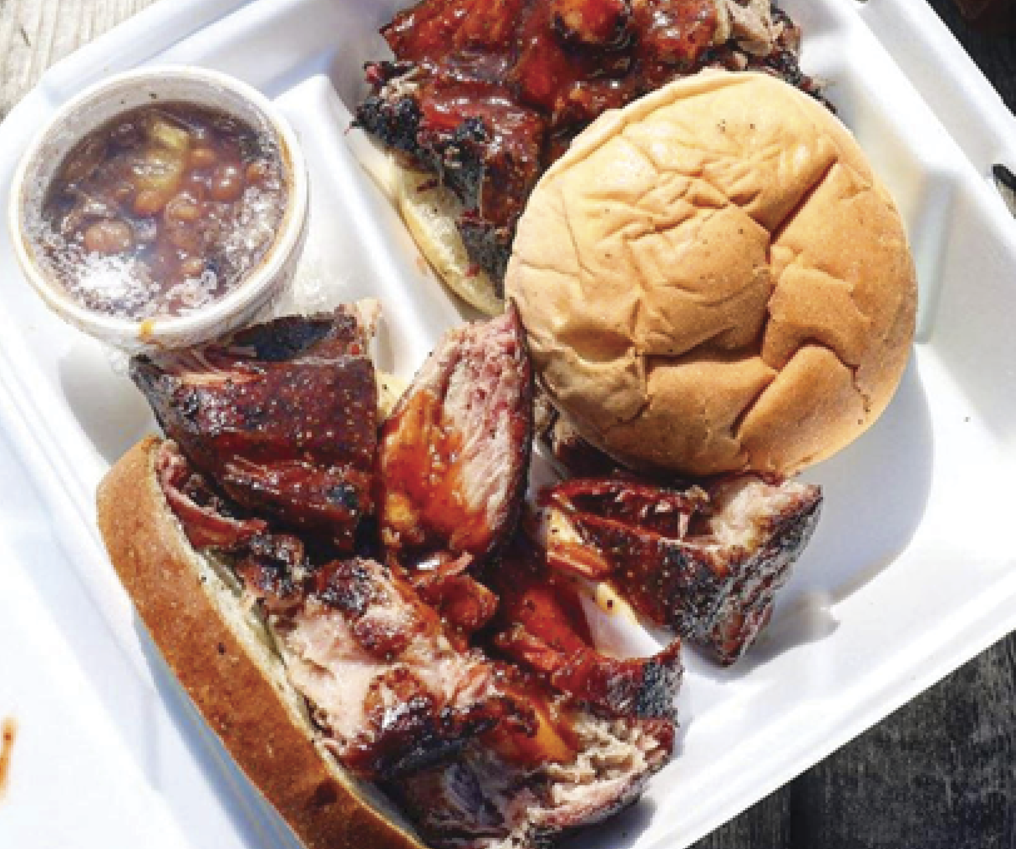 Top view of a white takeout plate with Jones Bar-B-Q’s burnt ends with a small container with sauce in it and a bun