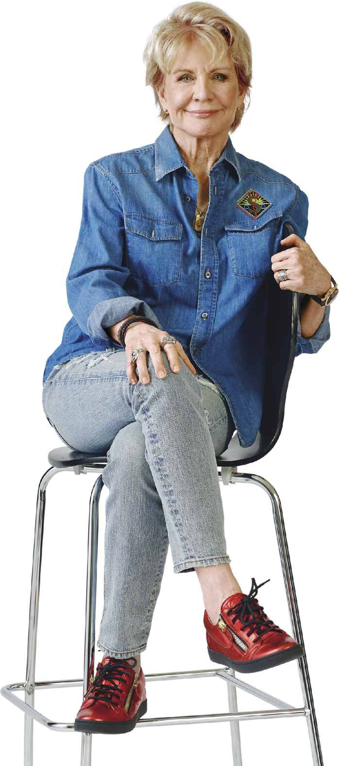 Image of Patricia Cornwell sitting on a barstool, looking into the camera and smiling. She is wearing a blue denim button-down shirt, jeans and red sneakers
