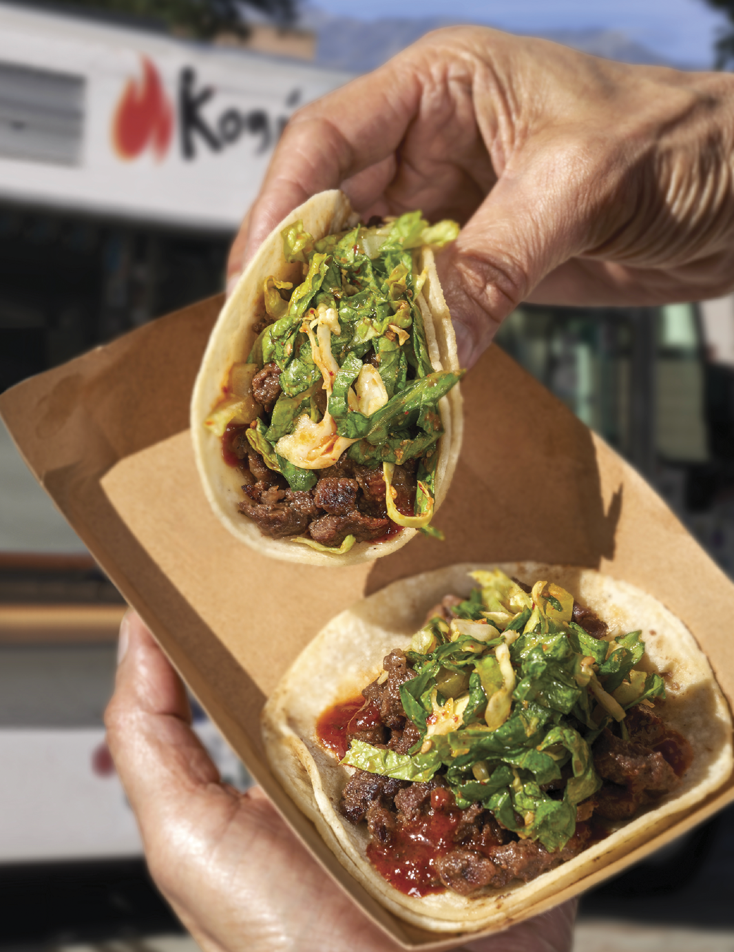 Hands holding a takeaway paper plate with 2 tacos in it. The tacos have a layer of meat topped up with a layer of seasoned greens