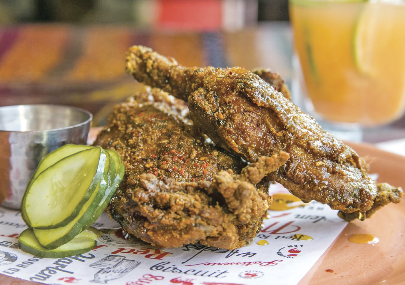 Image of Red Rooster’s fried chicken sitting next to a steel cup with a few slices of pickles resting on the fried chicken