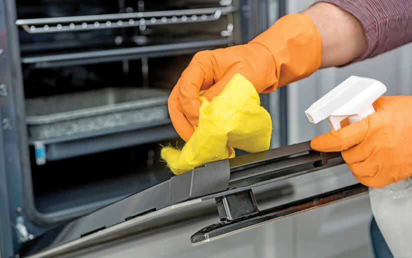 Photo of gloved hands cleaning an oven door with a rag and spray cleaner