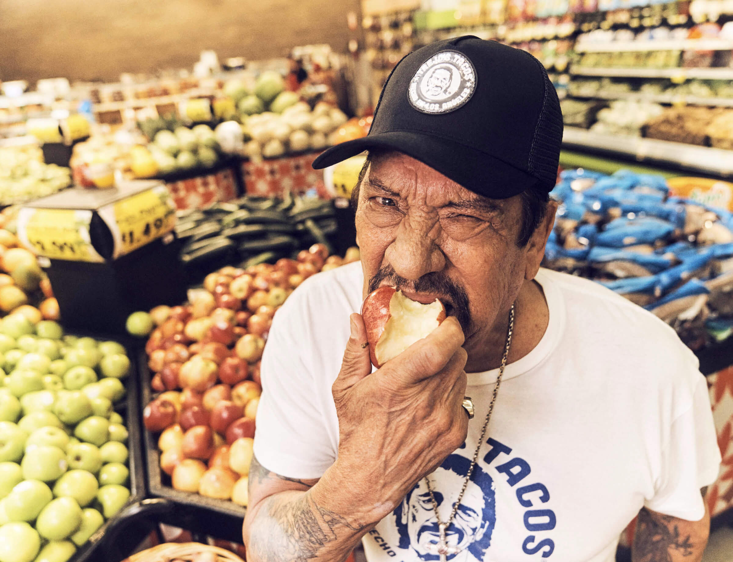 Photo of Danny Trejo taking a bite out of an apple in the produce section of a supermarket