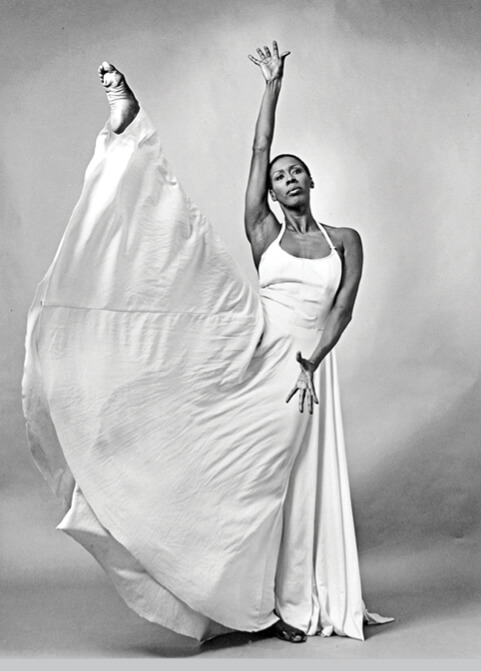 Photo of a younger Judith Jamison wearing a long white gown with her right leg and arm raised high