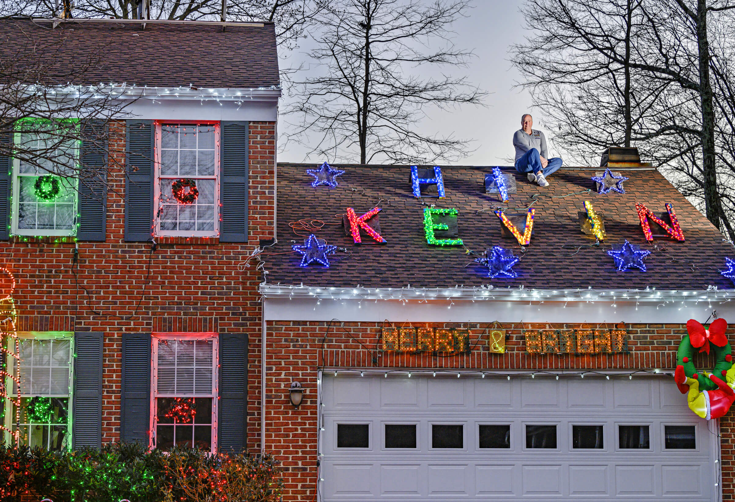 Photo of Mike Witmer sitting on the garage roof of his house decorated with Christmas lights