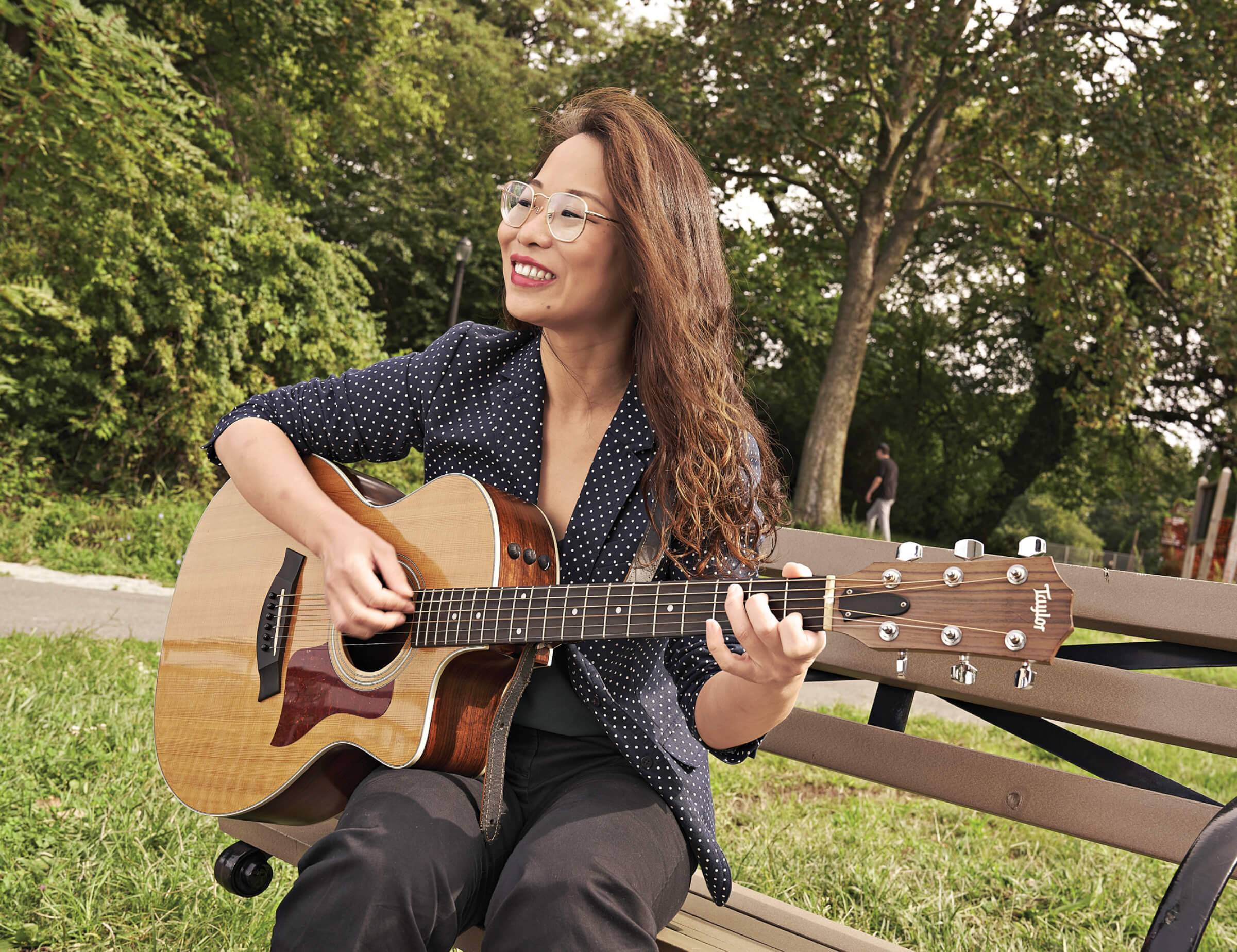 Photo of Xiyu Zhang playing her guitar while seated on a park bench