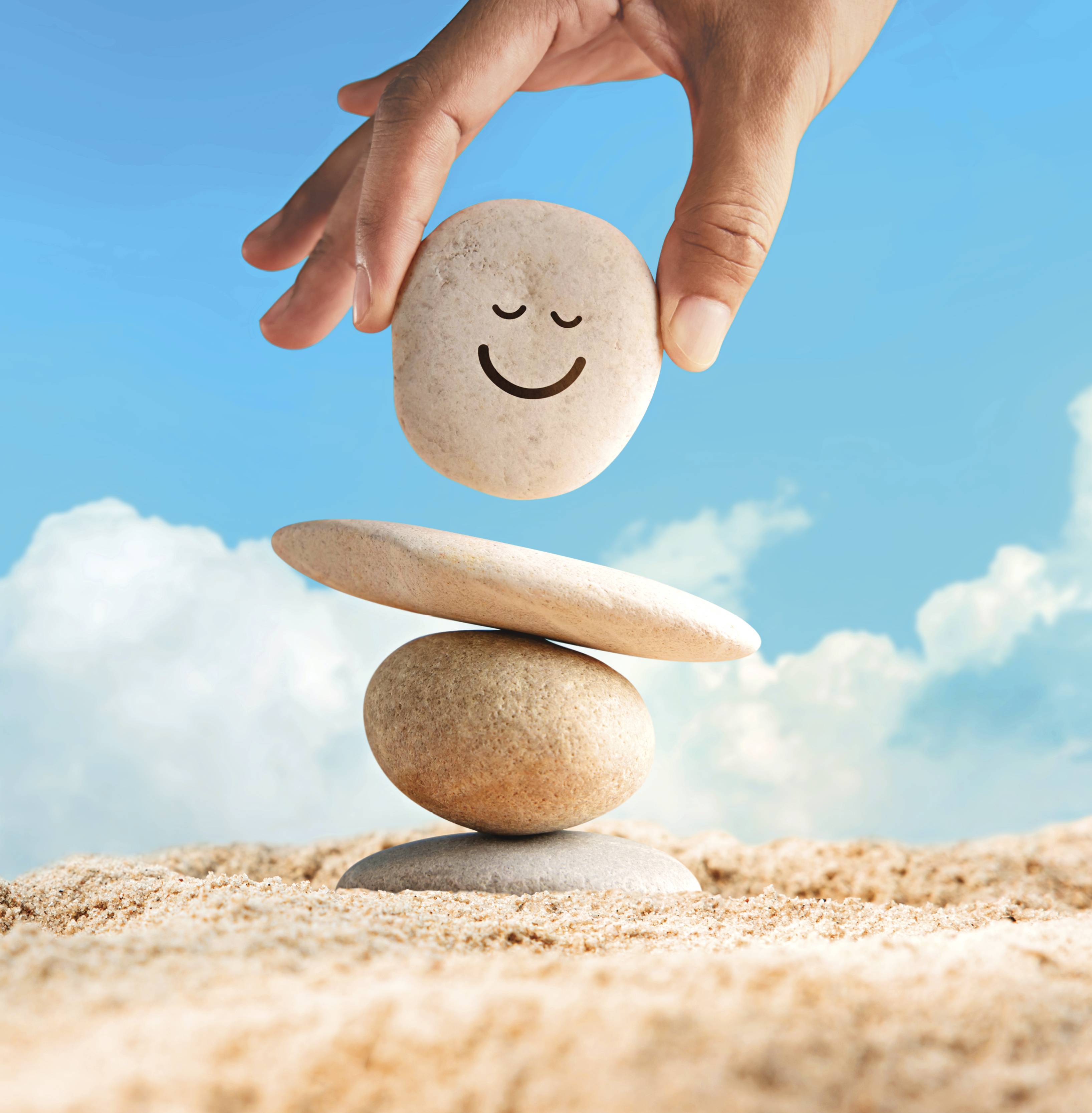 A close-up image of a human hand delicately balancing two smooth, round stones, one on top of the other, to form a small cairn. The topmost stone has black happy face drawn on its surface. 