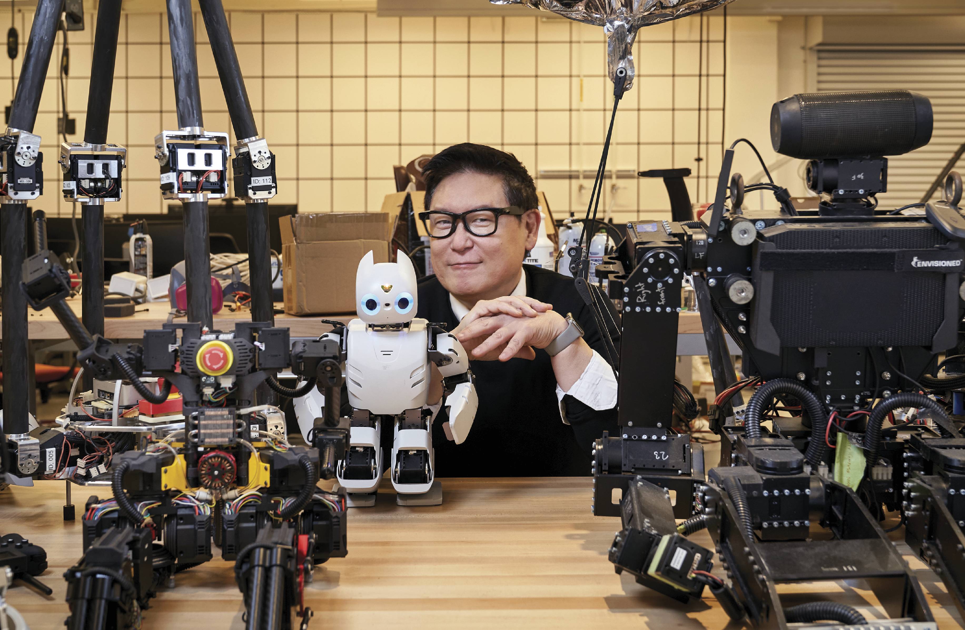 Photograph of Dennis Hong in his laboratory surrounded by several of his robotic creations, including a blue-eyed DARWIN