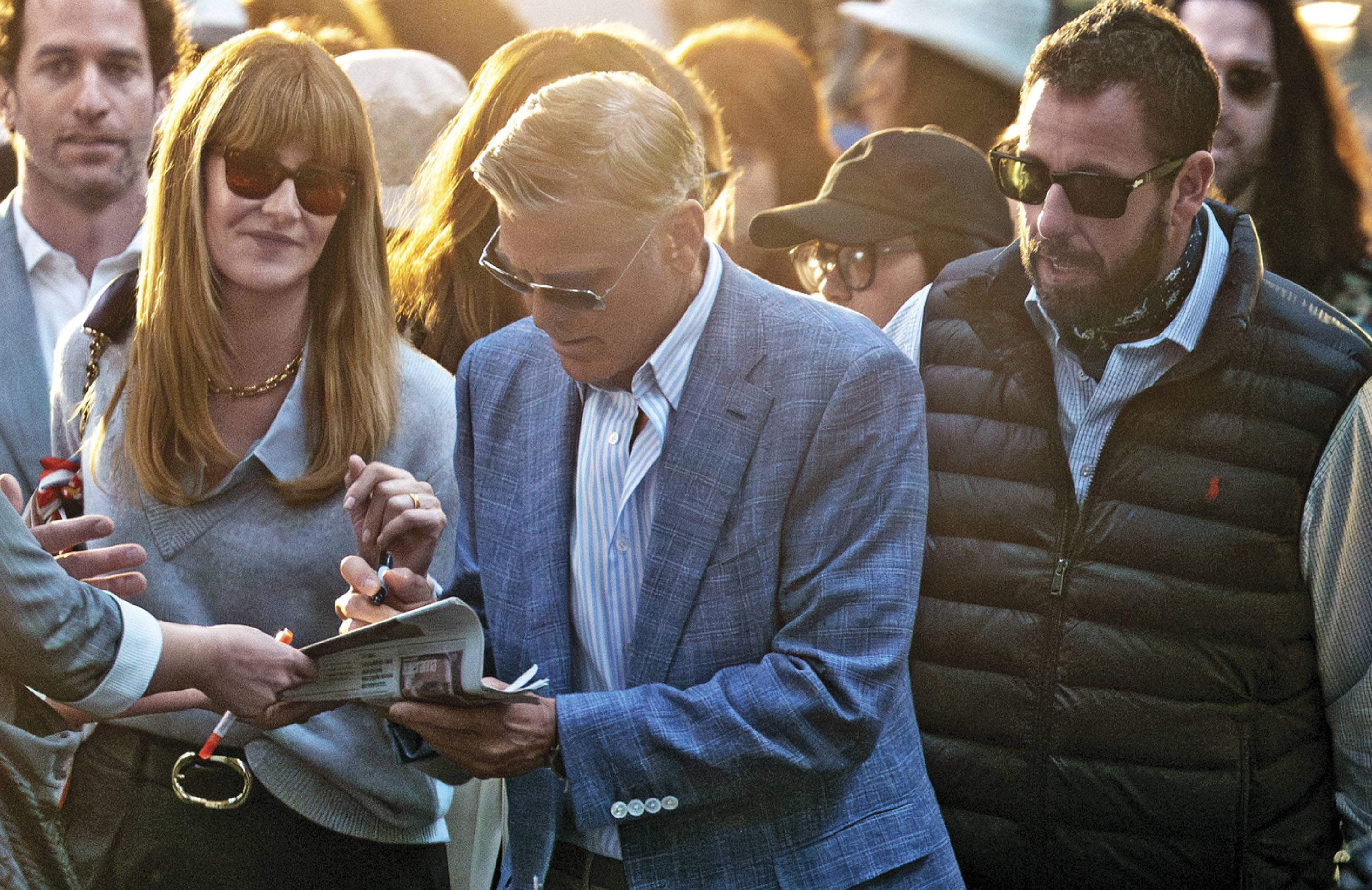 A group of people are gathered indoors. A man in the center wears a light suit and glasses, while a man to his right wears a dark quilted vest and sunglasses. The feature mentions actors George Clooney and Adam Sandler.