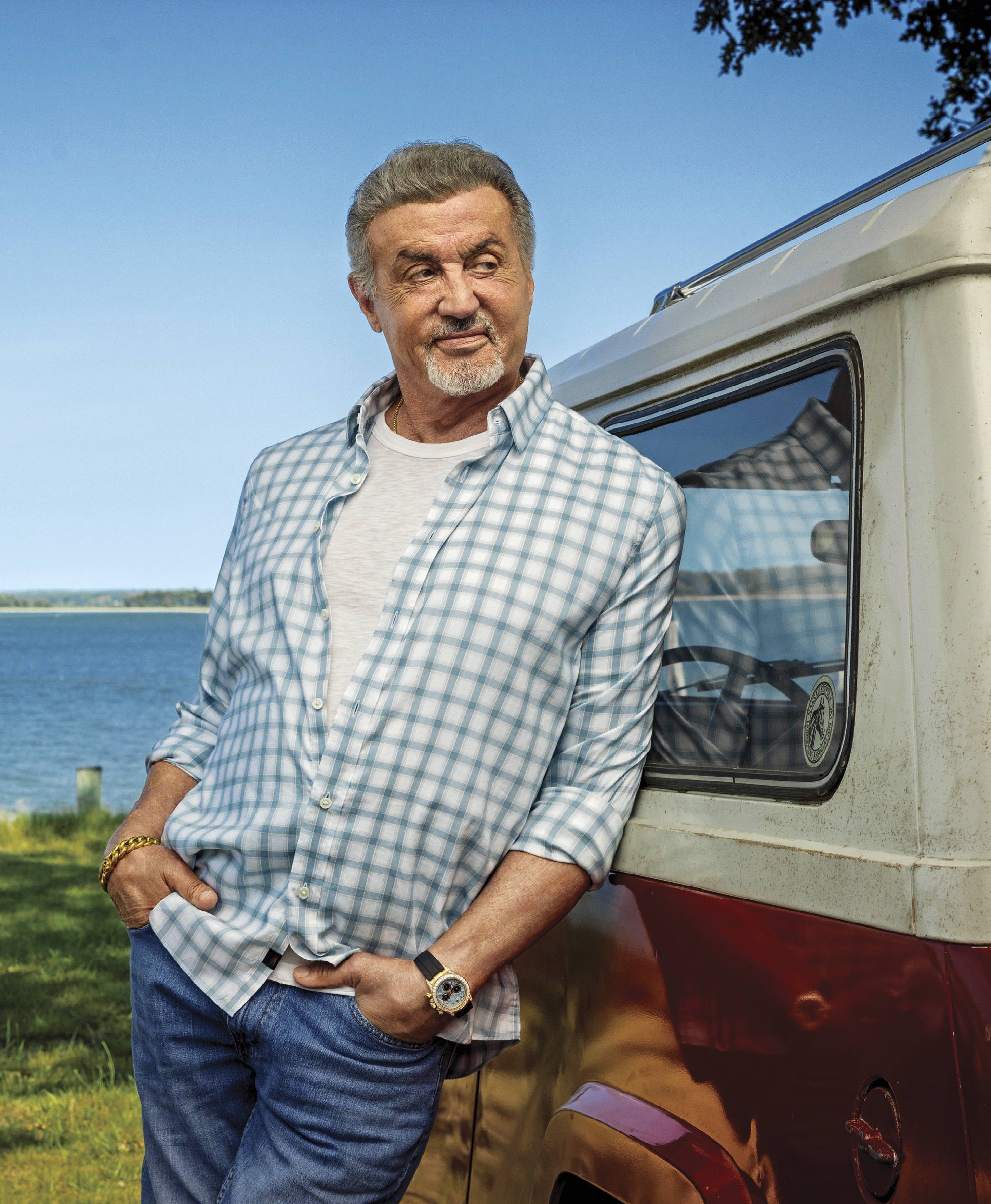 Image of Sylvester Stallone leaning against a trailer with blue water and greenery behind him 