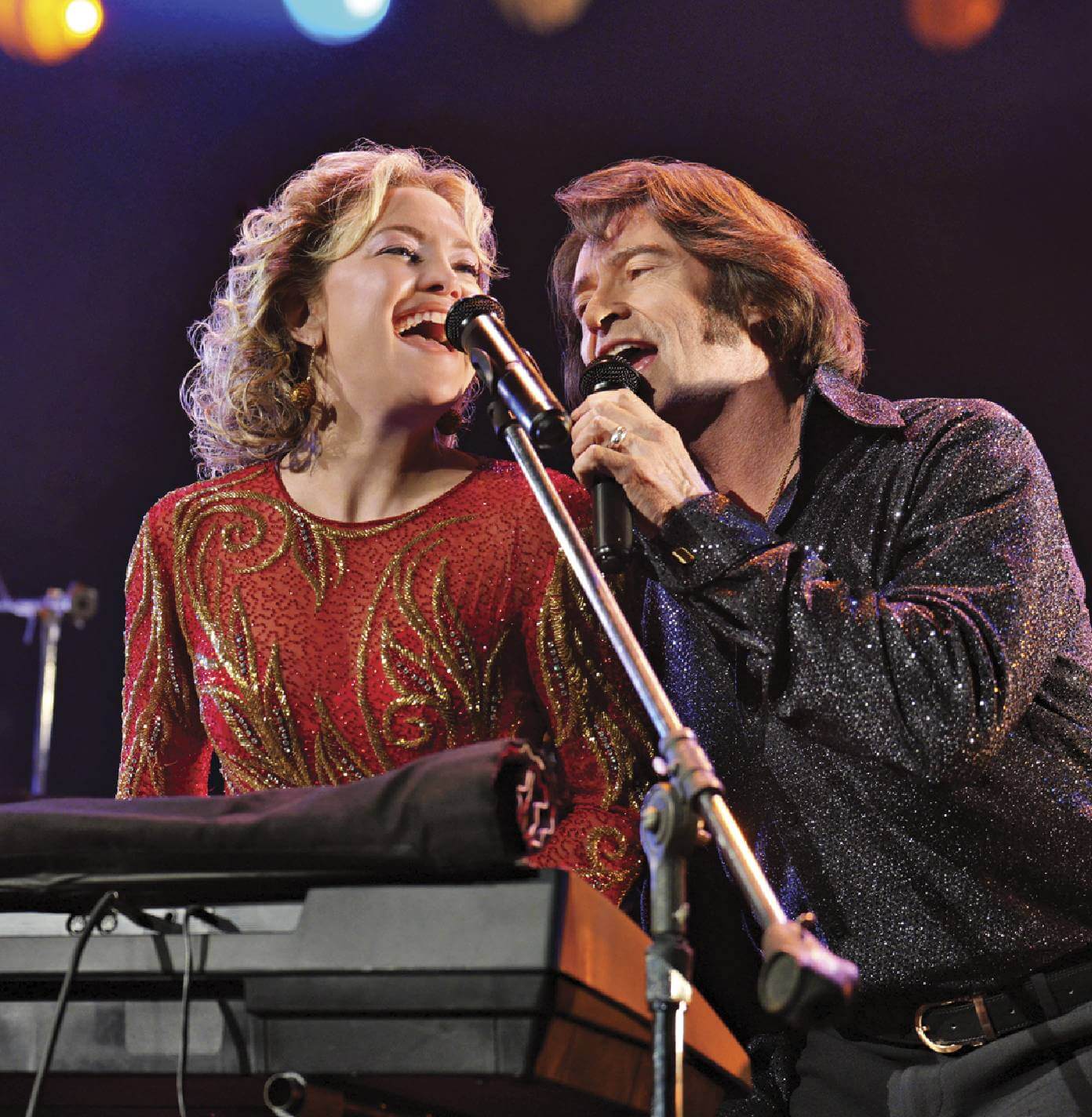 Two people are on a dark stage, leaning into microphones as if performing a musical number. The feature mentions Hugh Jackman (57) and Kate Hudson Star (50) in connection with the show. 