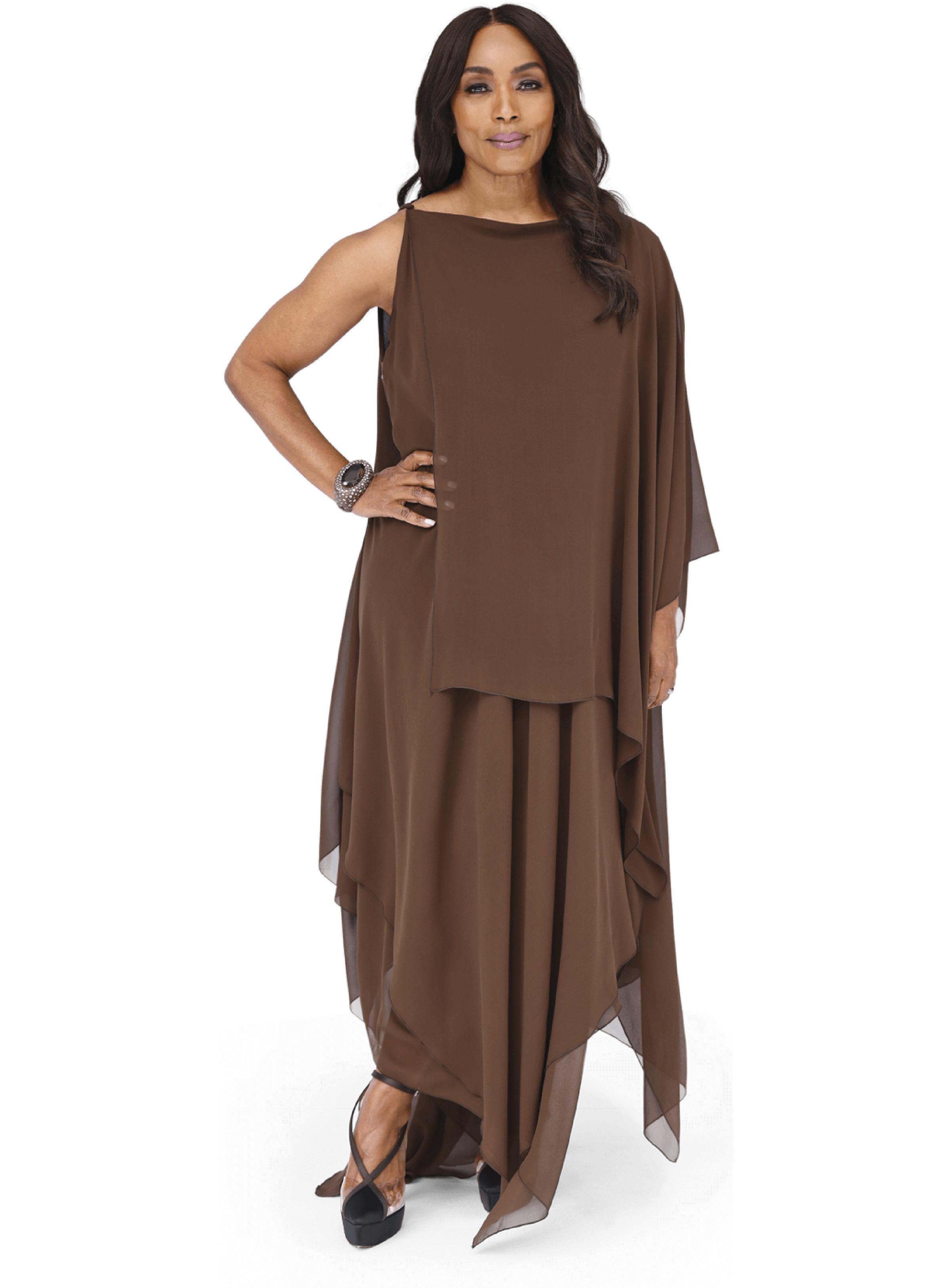 A full-body portrait of actor Angela Bassett standing against a solid beige background. She is wearing an elegant, flowing dress made of light brown/bronze fabric with a draped, asymmetrical design. She is looking directly at the camera with a calm expression, wearing subtle jewelry including a watch and rings.