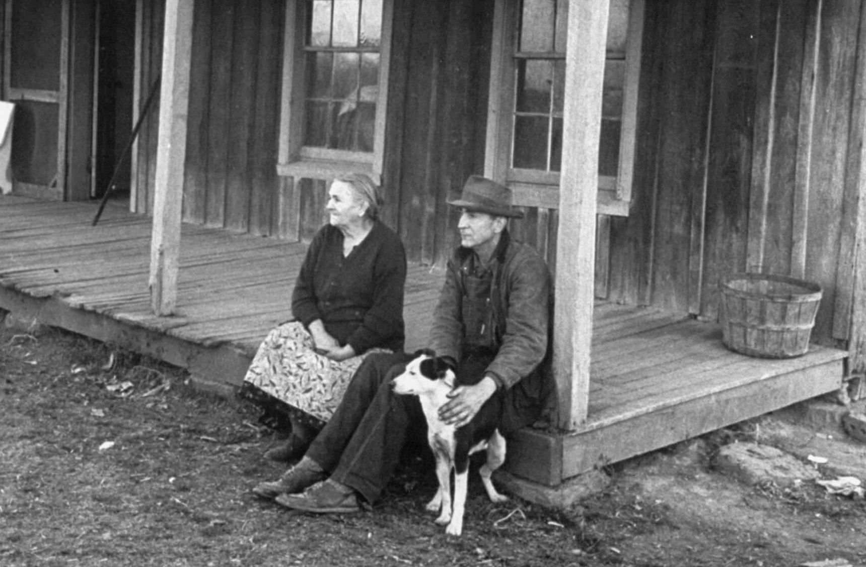 Black and white photo of a poverty-stricken couple sitting on the wooden porch of an old-looking house