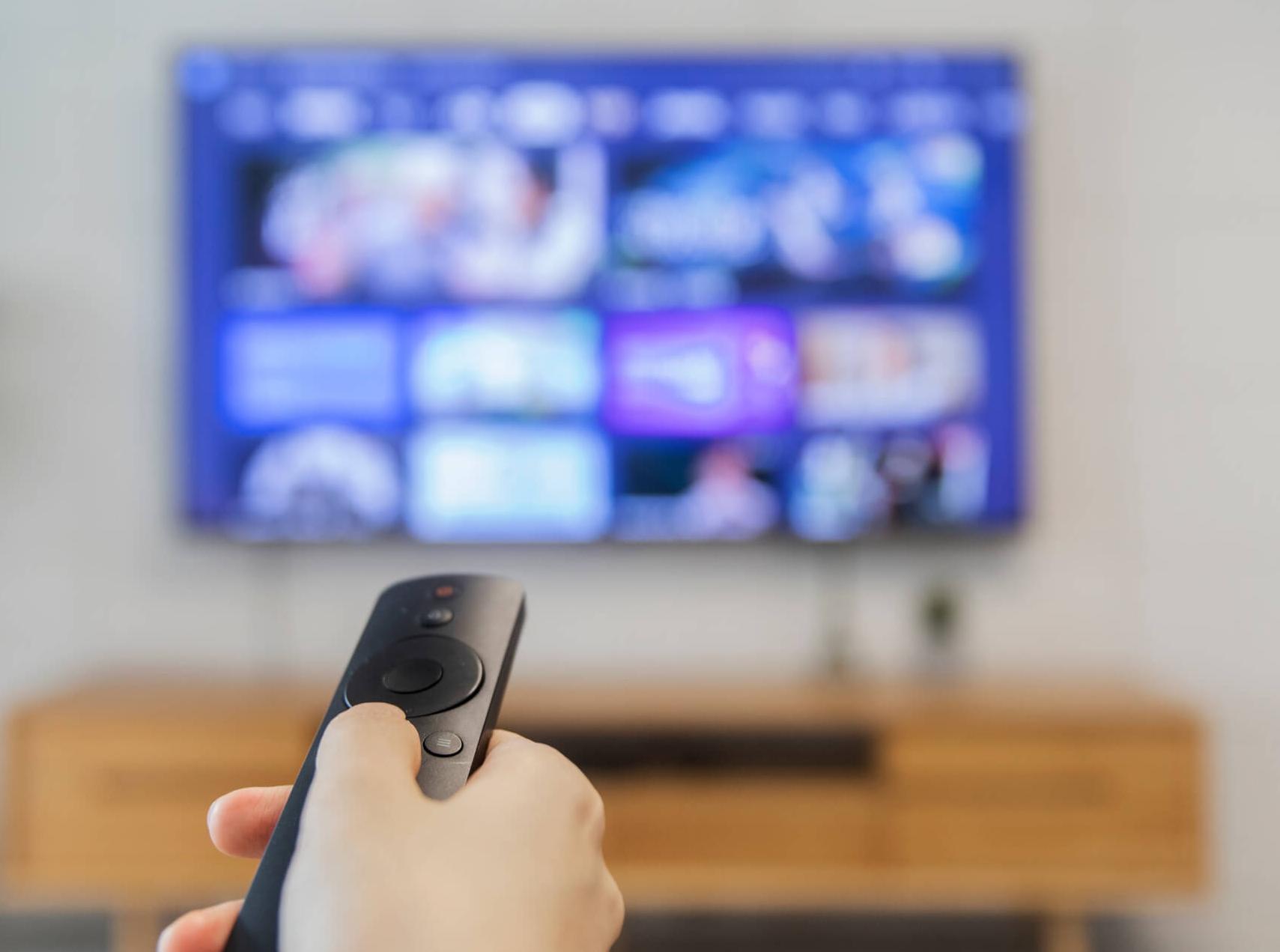 Close-up photo of a hand pointing a TV remote contral at a large flatscreen TV on the wall