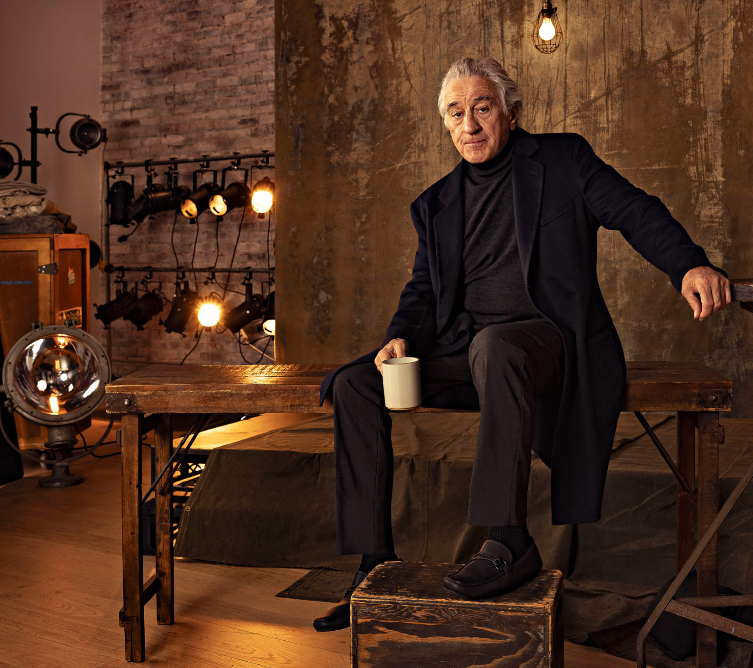 Photo of Robert De Niro dressed in black sitting on a wooden table