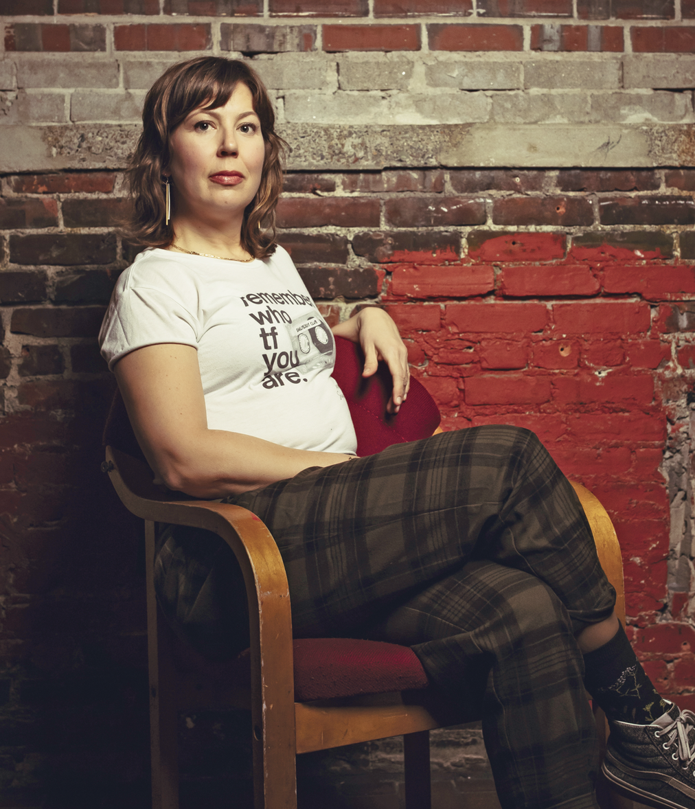 Woman in a white t-shirt and plaid pants sitting on a chair in front of a brick wall