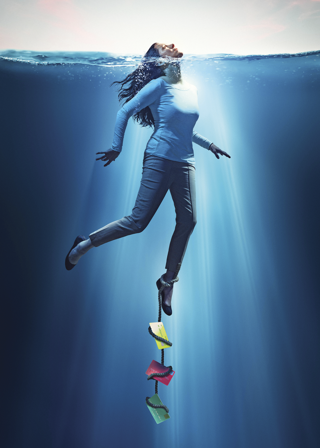 Woman underwater with only her head outside the water. A rope is tied to her leg while the other end is tied to several credit cards&nbsp;that are dragging her underwater
