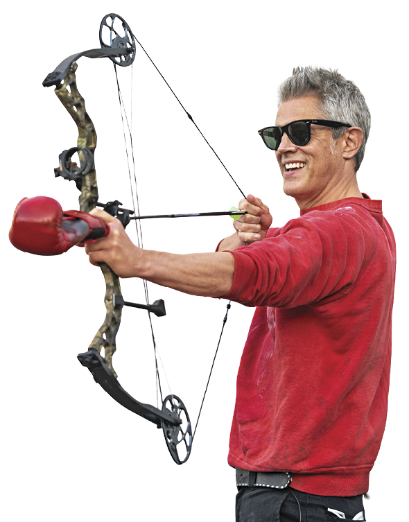 A man holding a bow and arrow. The arrowhead is a boxing glove and he is wearing a red shirt and sunglasses