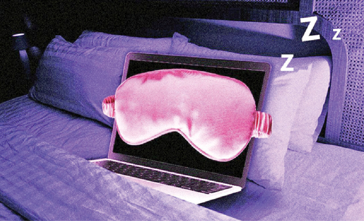 Photograph of a laptop open and resting on a bed with a pillow behind it. The laptop is wearing an eye mask and sleeping 