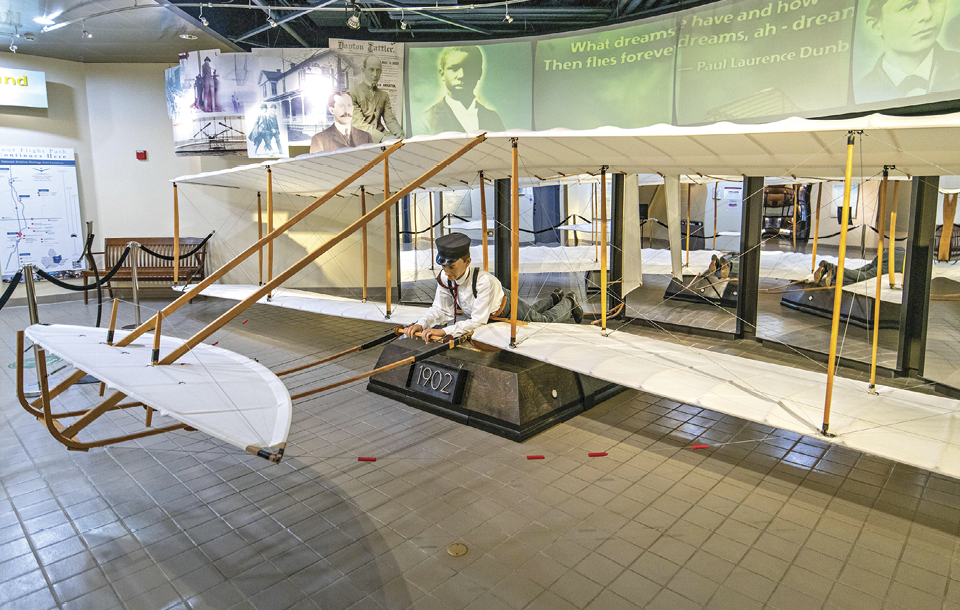 A full-scale replica of an early Wright brother’s aircraft is displayed on a museum floor, showcasing its intricate wooden frame and fabric wings.