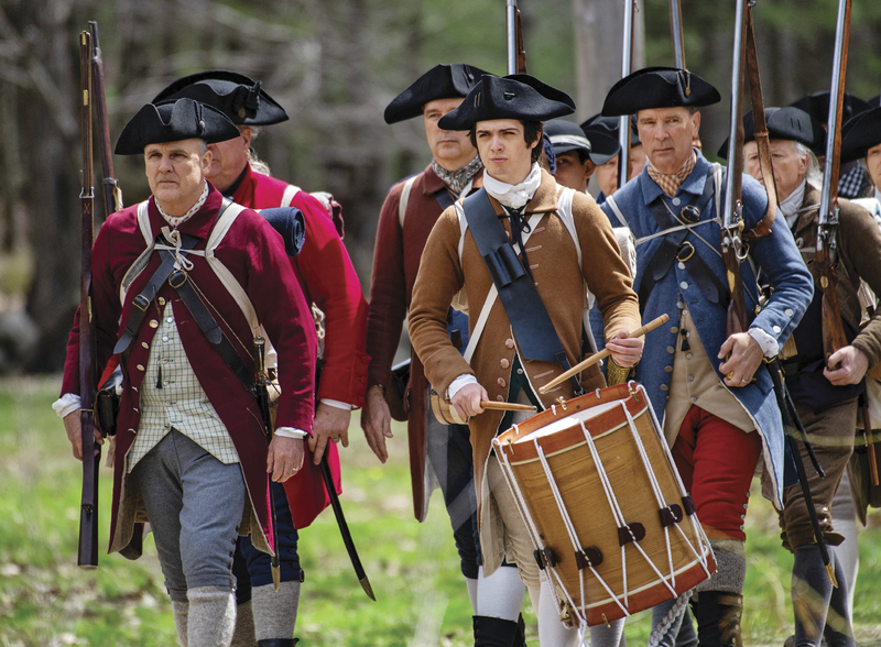 A group of reenactors marching outside, dressed in American Revolutionary War-era clothes. 